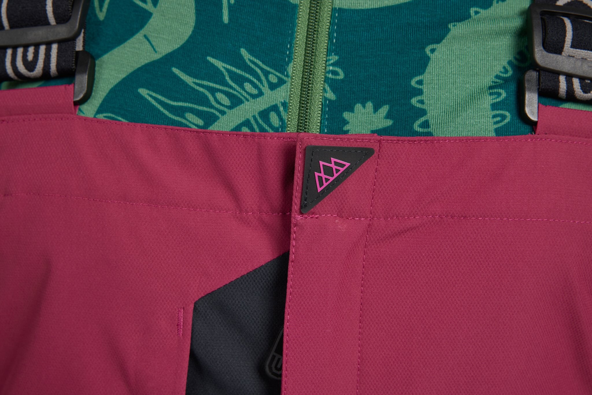 Close-up detail of a pink technical outdoor garment with a black triangular logo tab featuring a pink mountain symbol, showing part of a green patterned top underneath.