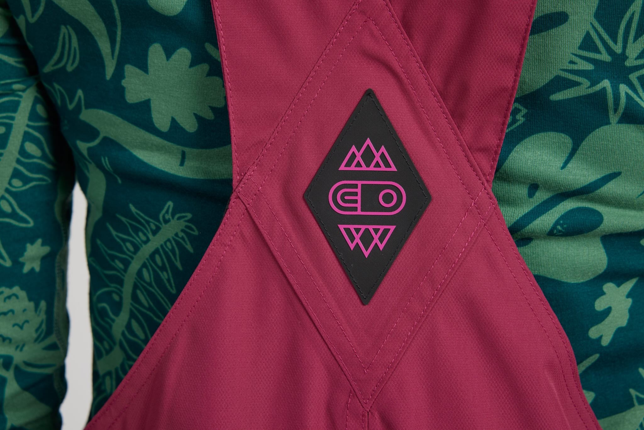 Close-up of a pink bib-style garment with a black diamond patch featuring a pink geometric logo, worn over a green tropical-patterned long-sleeve top.