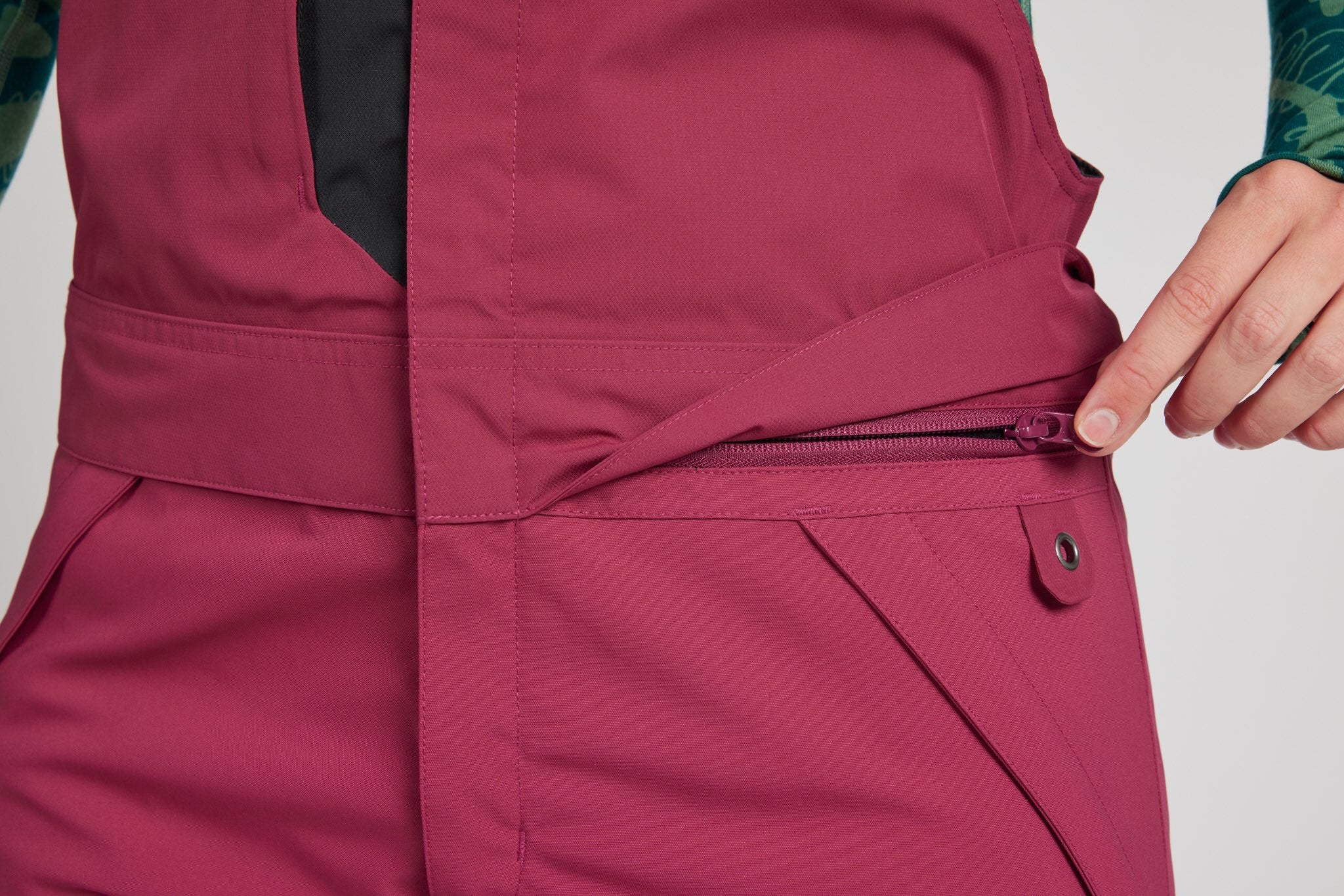 Close-up of a person wearing a dusty rose pink technical snowboard bib, showing a zippered pocket and a hand adjusting the strap.