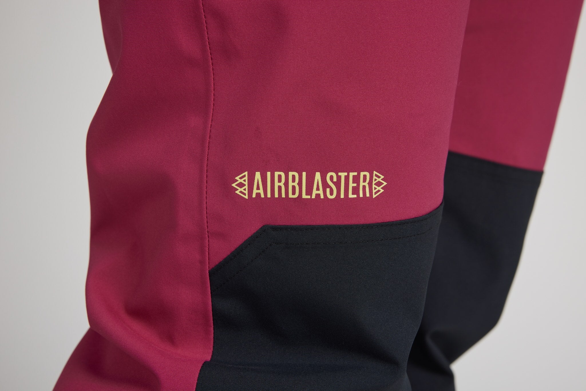 Close-up of a ski jacket leg showing a colorblock design with a magenta upper section, black lower leg panel, and a gold AIRBLASTER logo on the side.