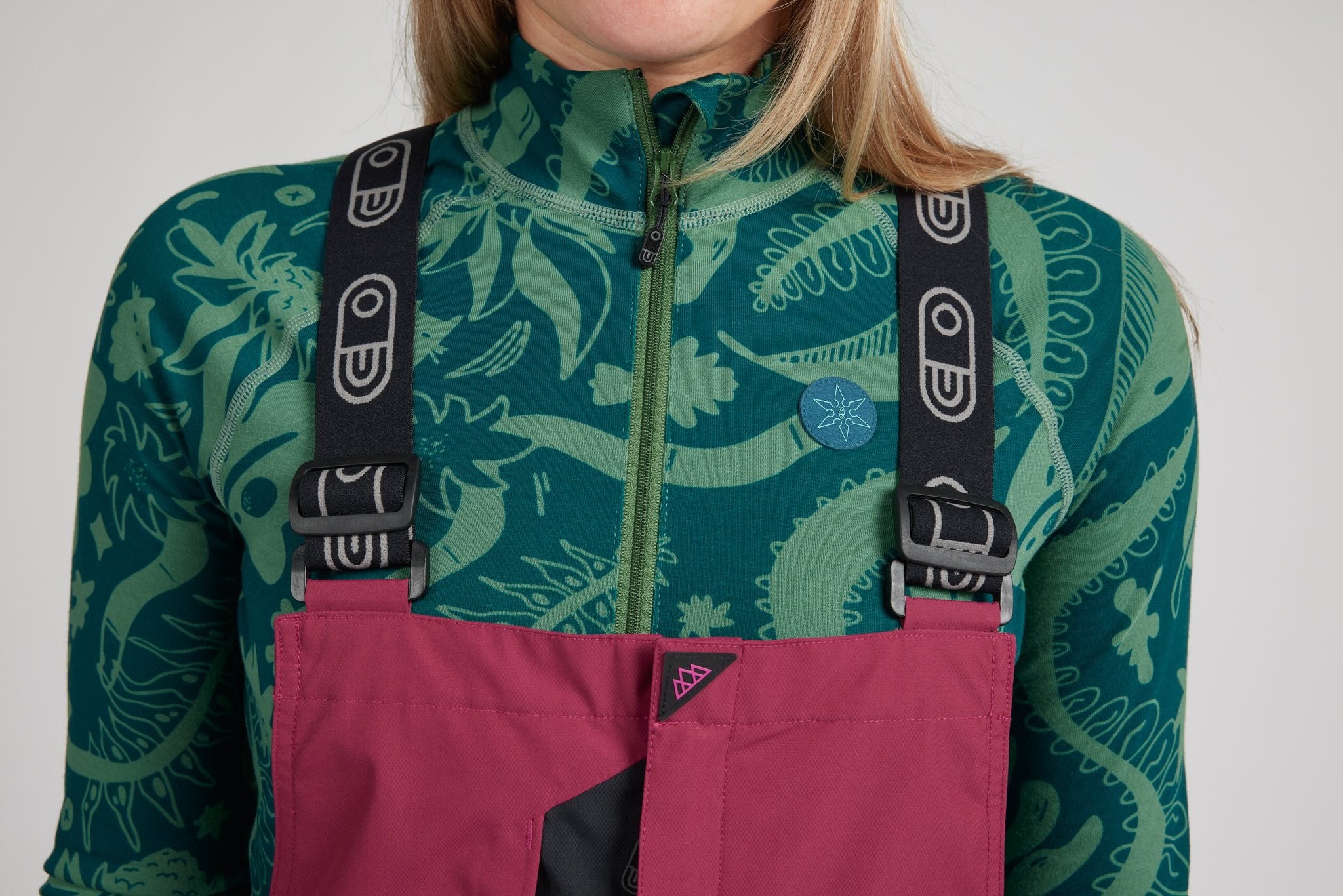 A close-up of a person wearing pink and black bib overalls over a green patterned quarter-zip fleece. The overalls feature a magenta-pink body with black adjustable straps and black plastic buckles.