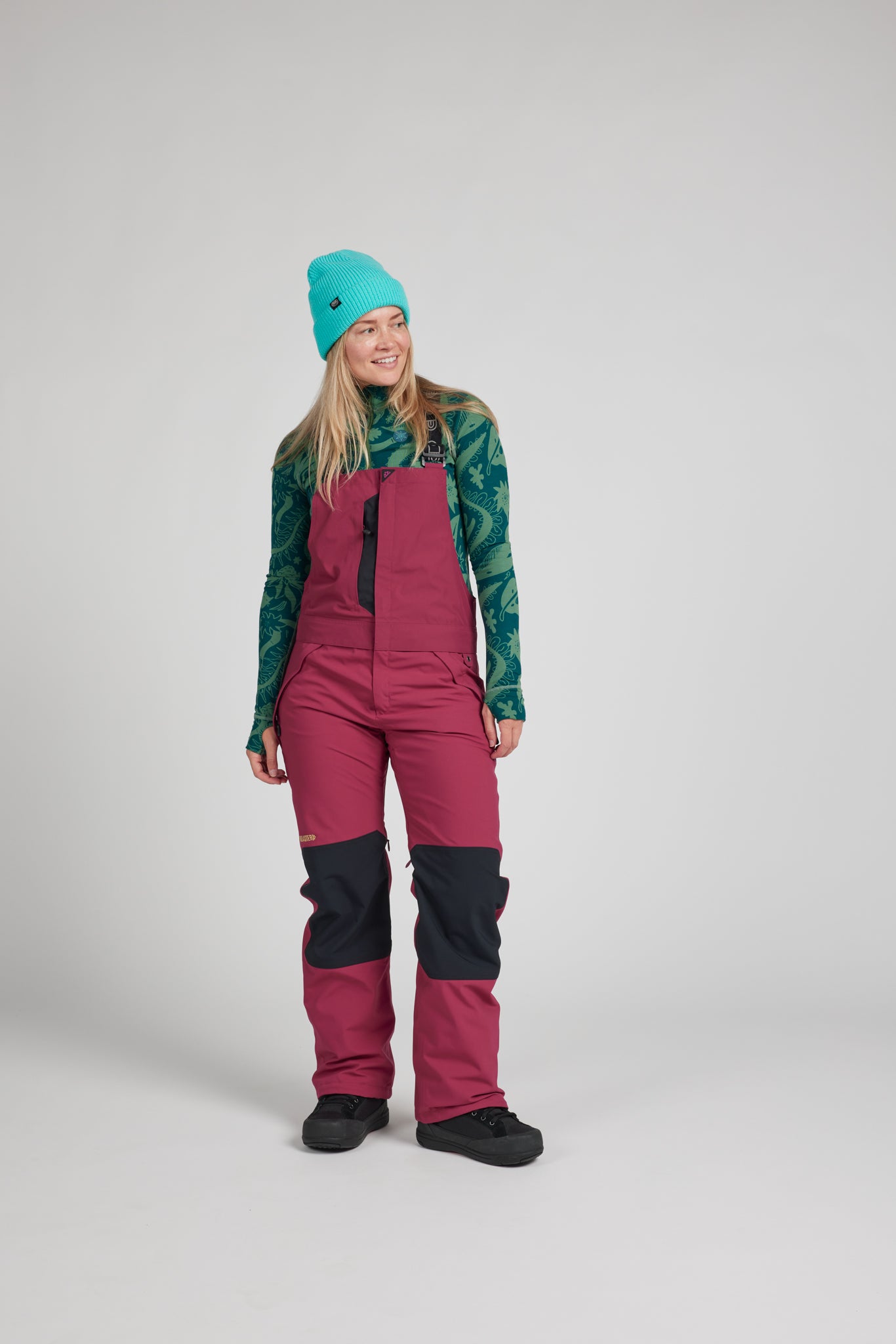 A woman modeling a pair of ski bib pants in a dusty rose and black colorway, paired with a green patterned long-sleeve top