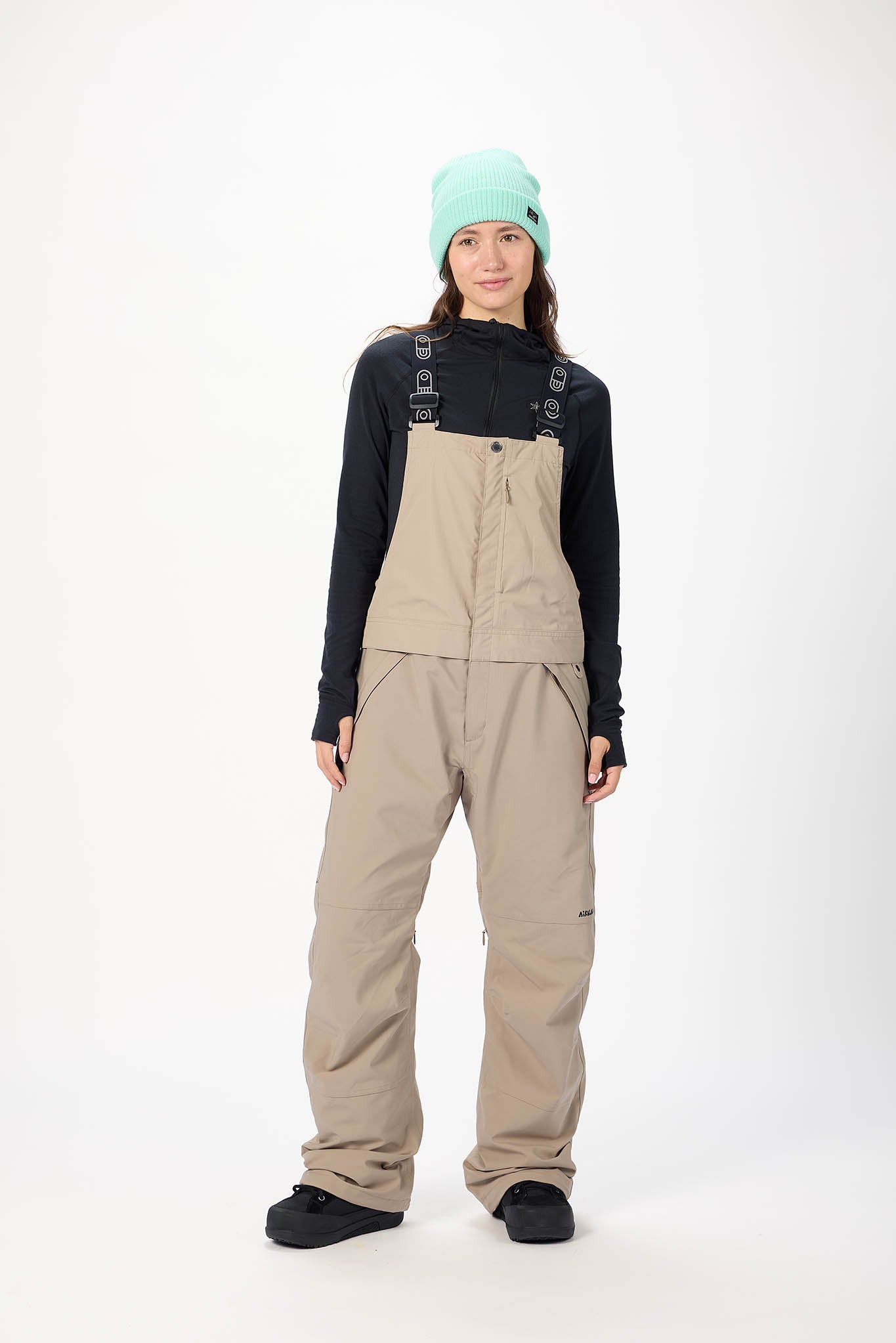 Person wearing a black jacket, beige bib overalls, and a light green beanie on a white background
