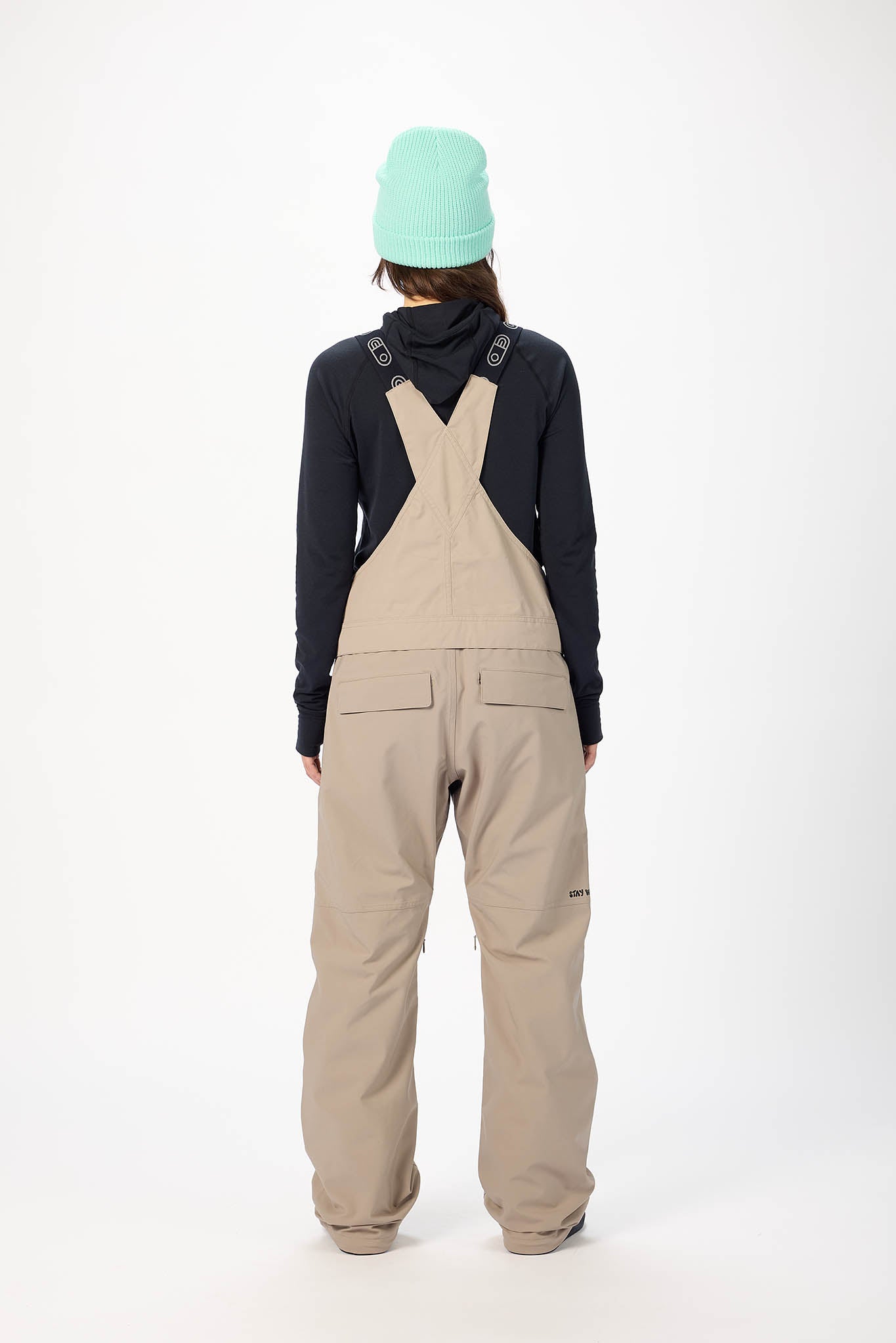 Person wearing a navy jacket, beige bib overalls, and a light green beanie on a white background