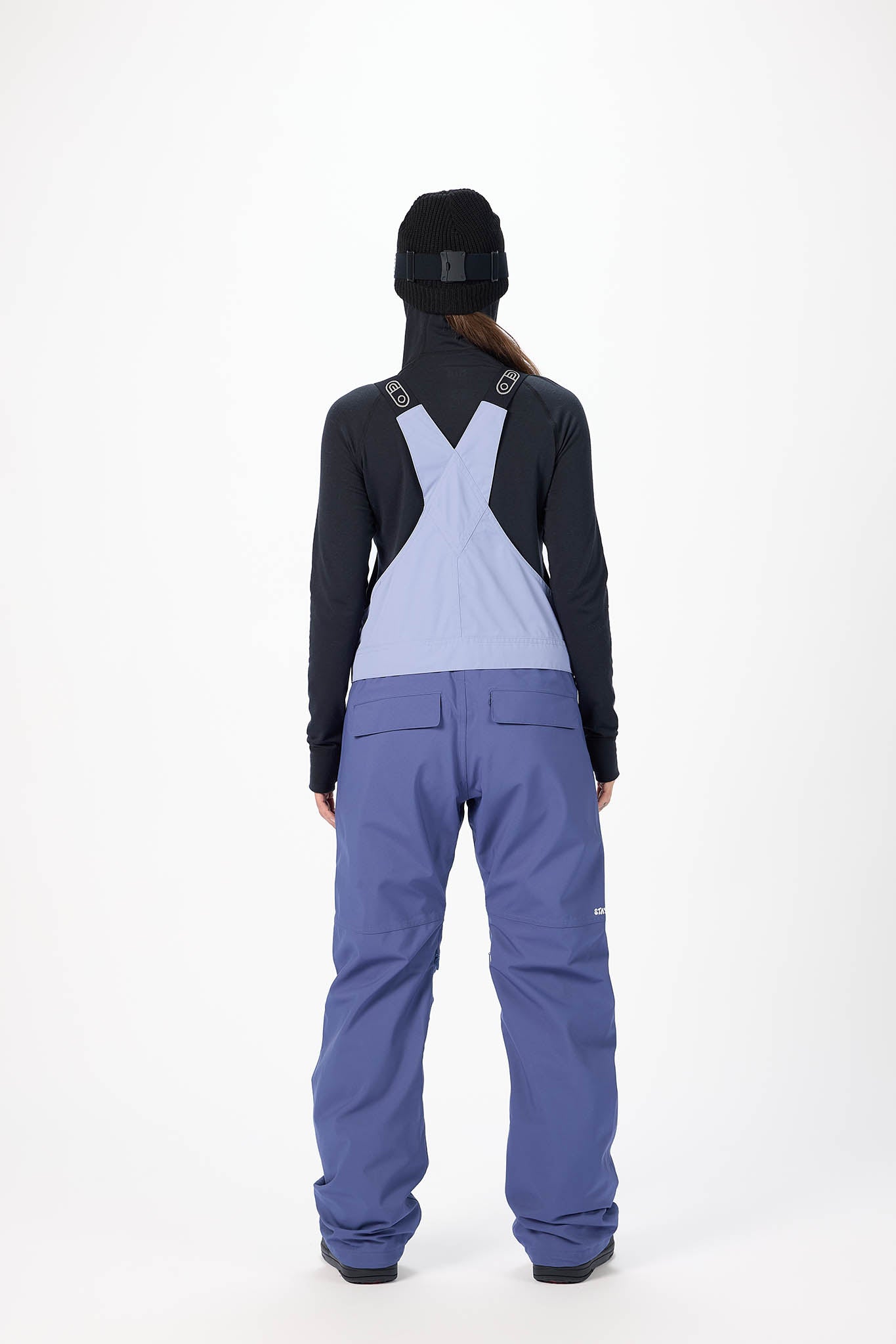Person wearing a black jacket and blue snow pants with suspenders on a white background
