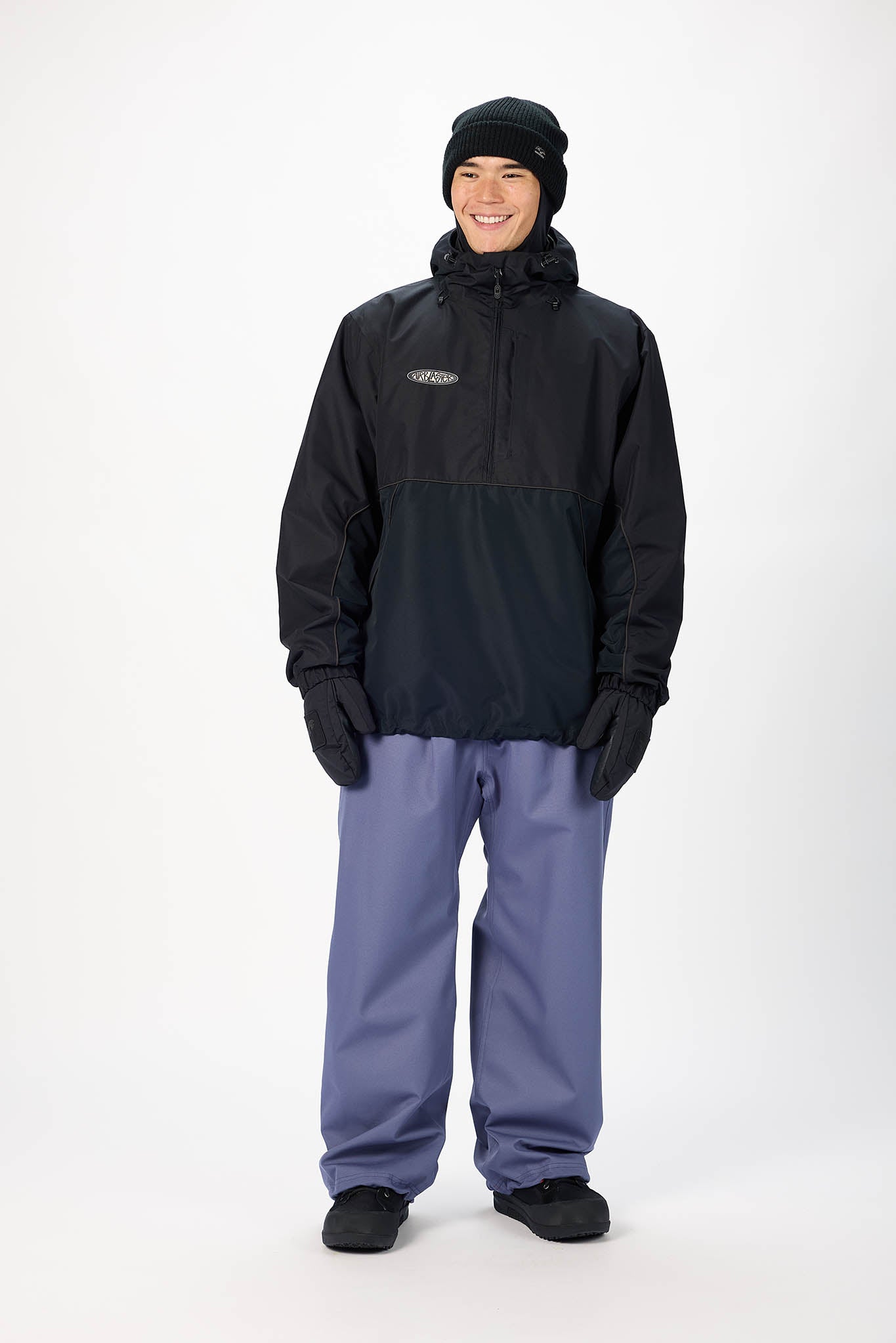 A man modeling a black half-zip hooded jacket, black beanie, black gloves, and slate blue snow pants against a plain white studio background.
