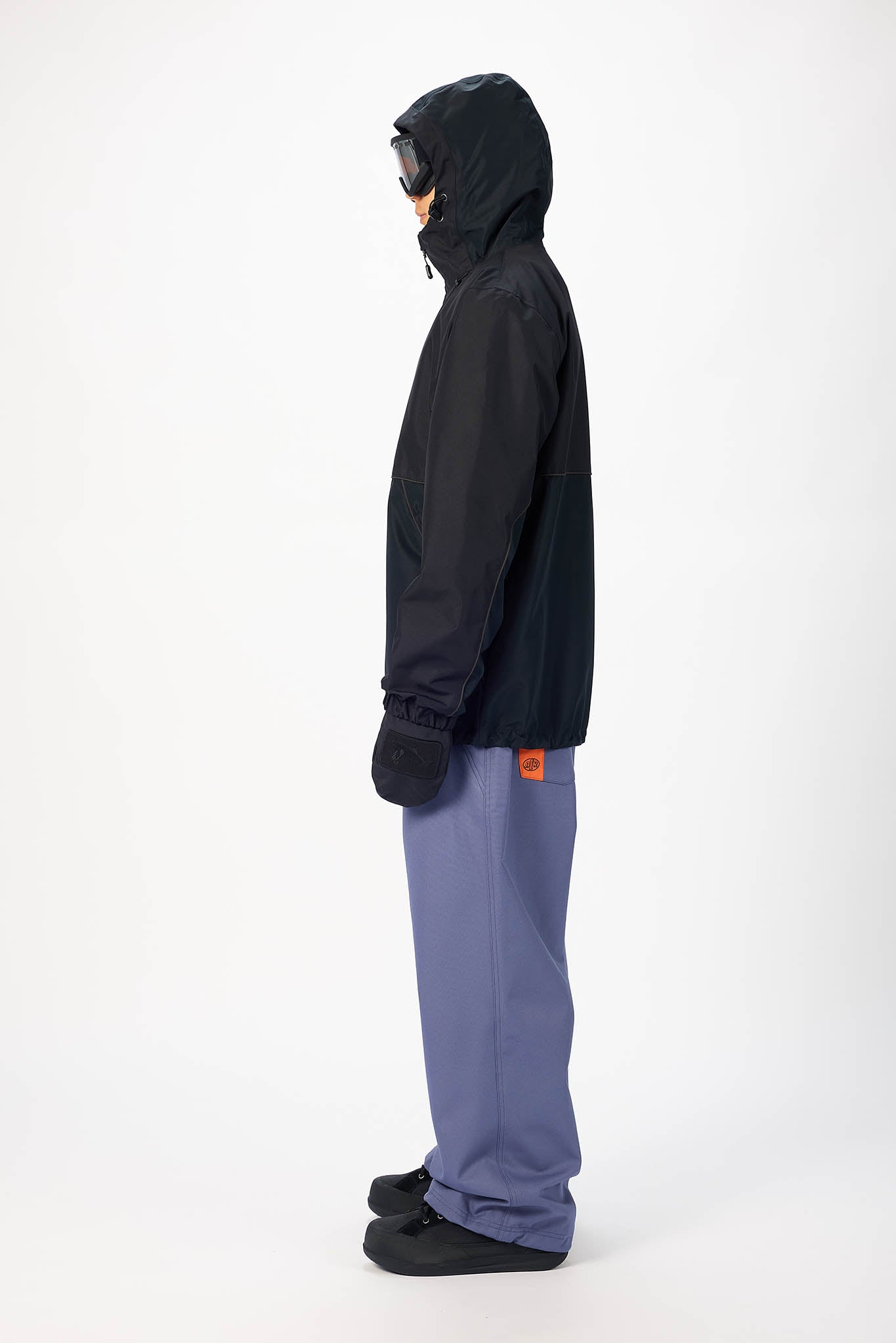 A model shown in profile wearing a black hooded jacket, slate blue pants, and black gloves against a plain white studio background.