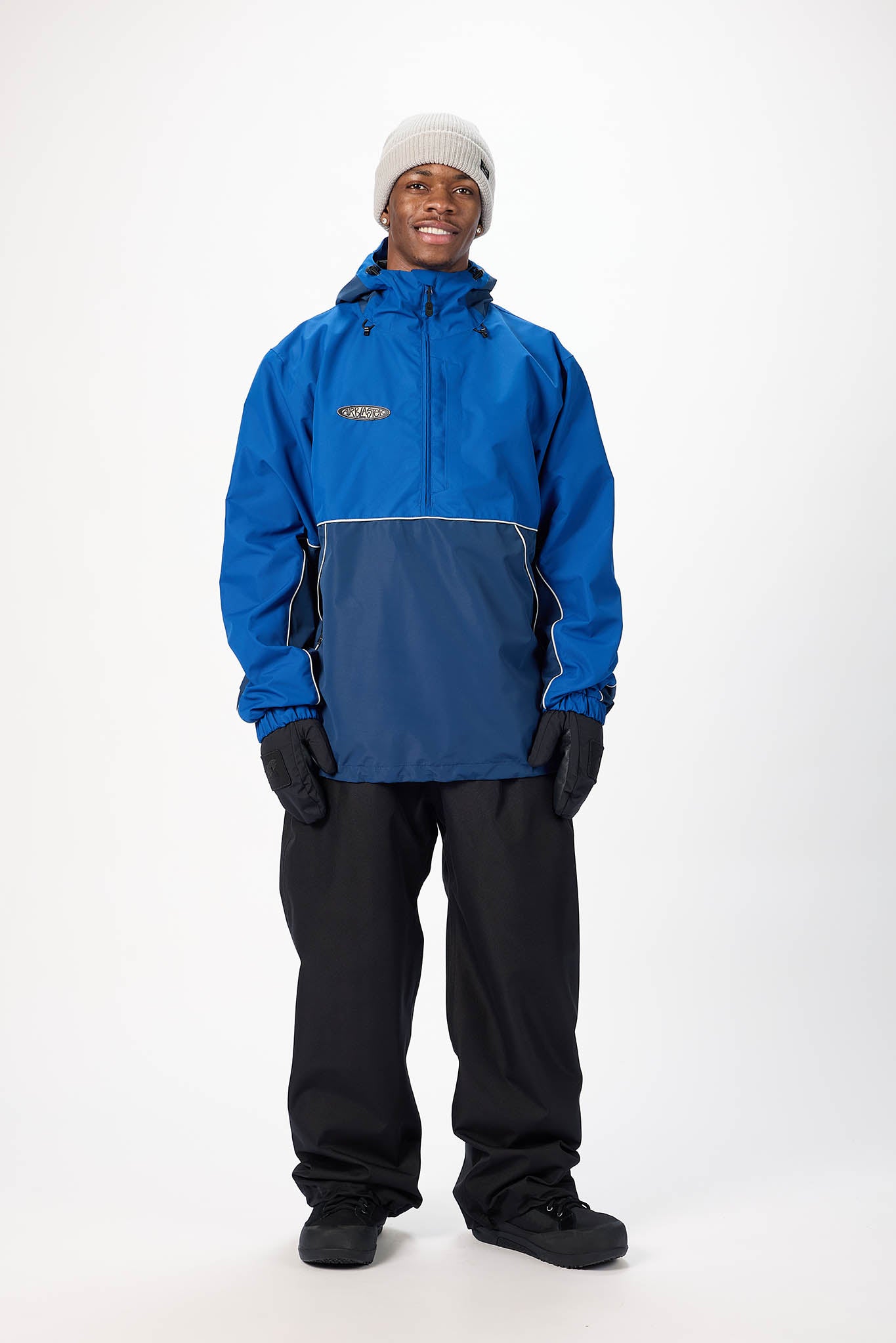 A man wearing a blue and navy colorblock snowboard jacket, black snow pants, black gloves, and a light gray beanie, standing against a white studio background.