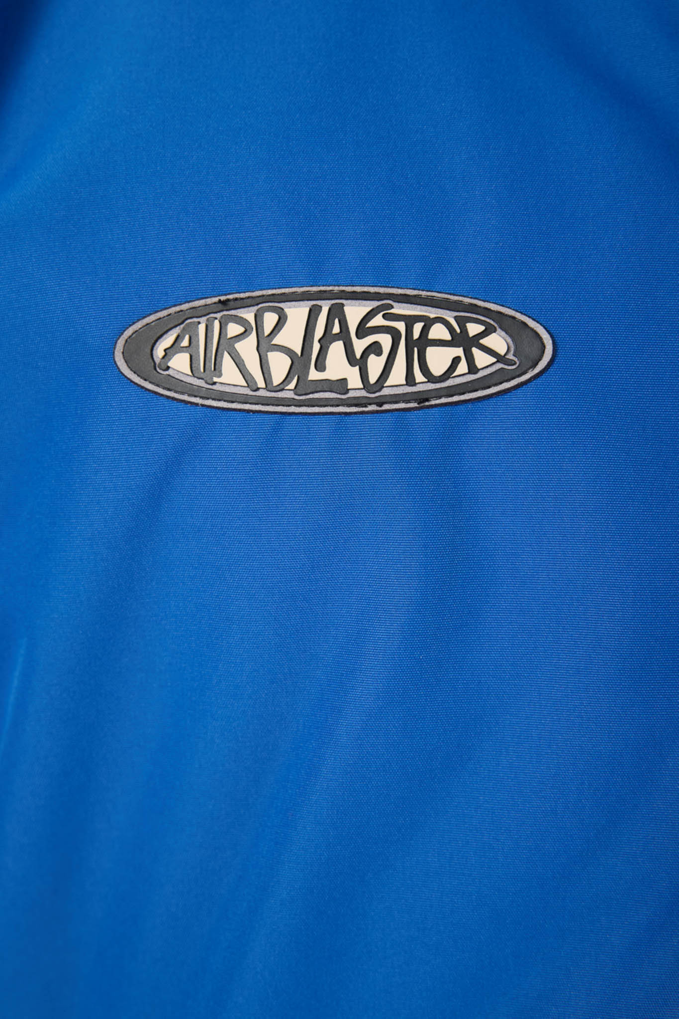 A close-up of a bright blue garment featuring an oval embroidered patch with the word 'AIRBLASTER' in black script on a white background with a dark border.