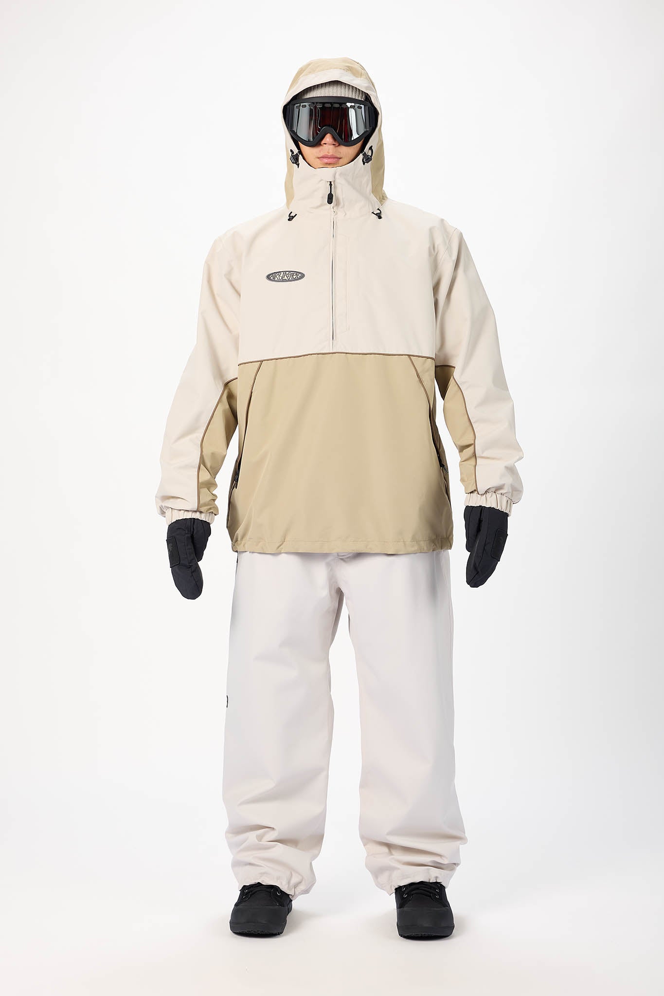 A model wearing a beige and khaki color-blocked snowboard jacket, white snow pants, black gloves, and goggles, standing against a plain white studio background.