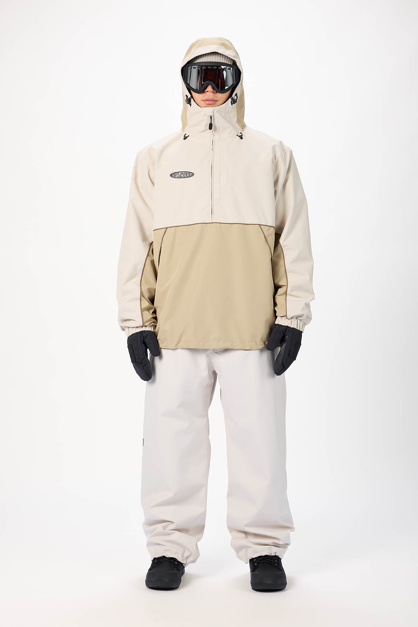 A model wearing a beige and khaki color-blocked snowboard jacket, white snow pants, black gloves, and goggles, standing against a plain white studio background.