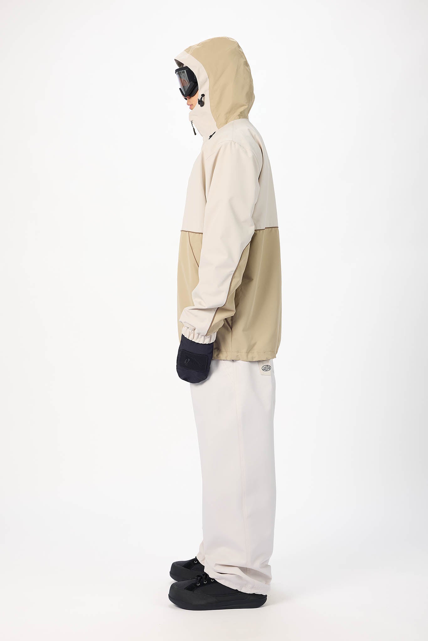 A person shown in side profile wearing a hooded jacket with a beige and off-white color-block design, off-white pants, black gloves, and black shoes, photographed against a plain white studio background.