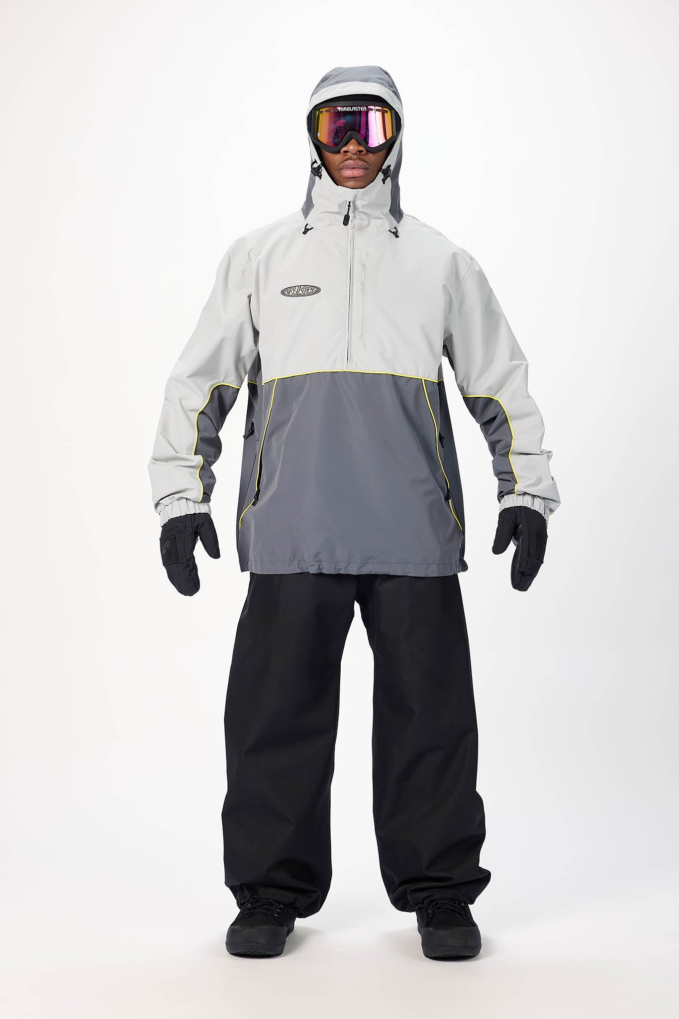 A model wearing a colorblock ski jacket in light gray and dark gray, paired with black snow pants, black gloves, and ski goggles, standing against a plain white studio background.