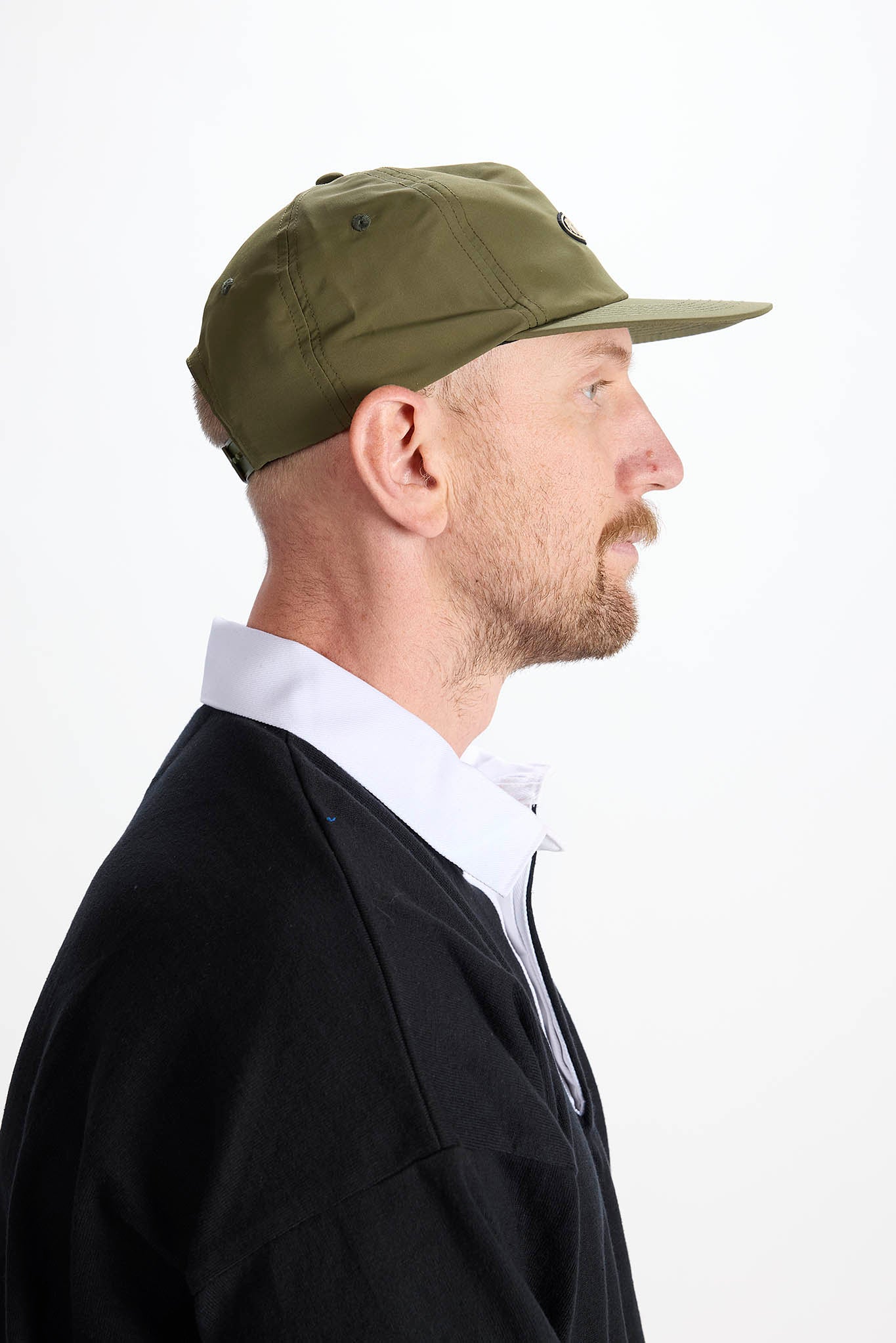 side profile A man wearing an olive green baseball cap with a white embroidered logo and a black long-sleeve polo shirt over a white collared shirt, photographed against a plain white background.