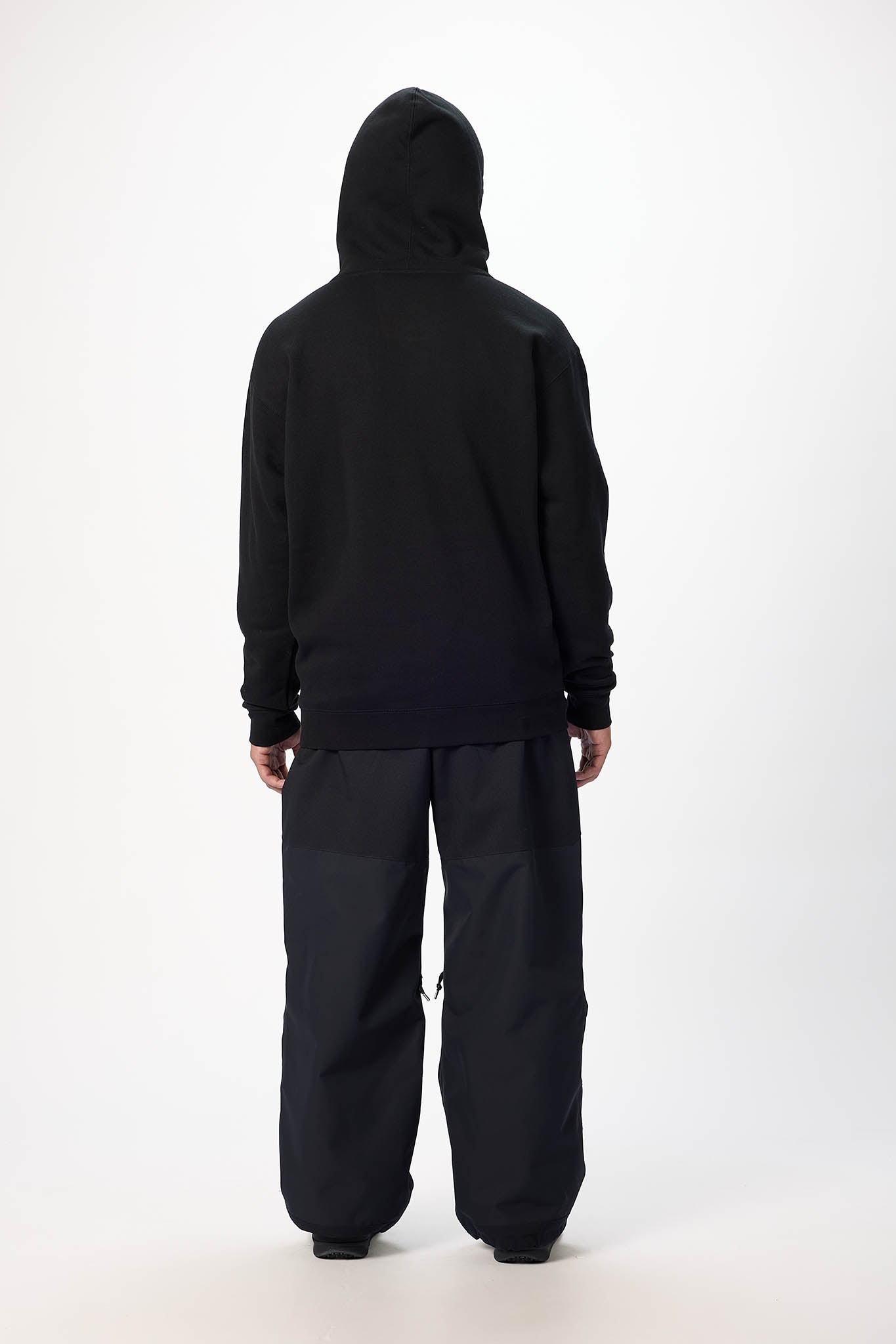 A person shown from the back wearing a black hooded sweatshirt and loose black pants against a plain light gray studio background.