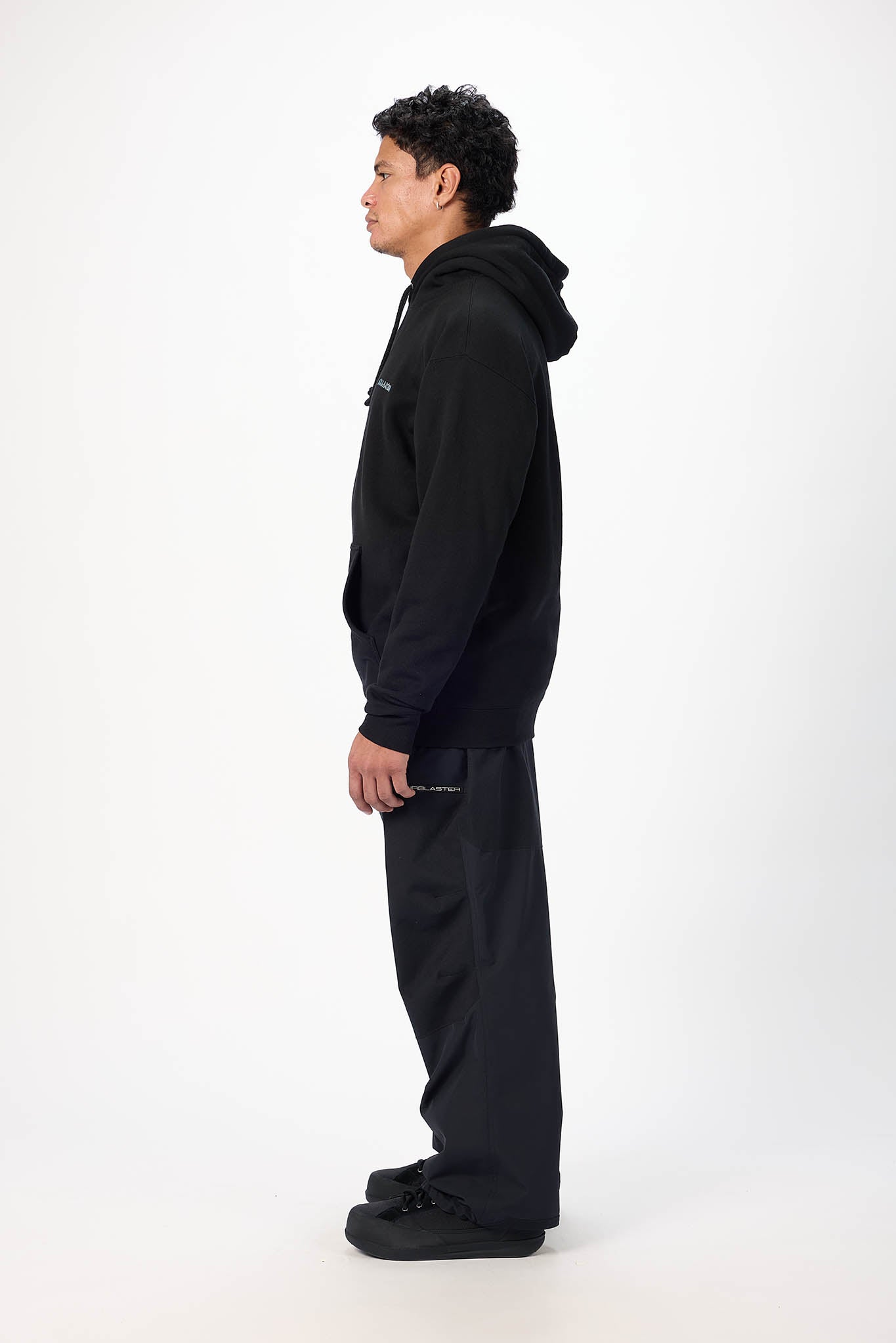 A man in a black hoodie and black pants stands in profile against a plain white studio background.