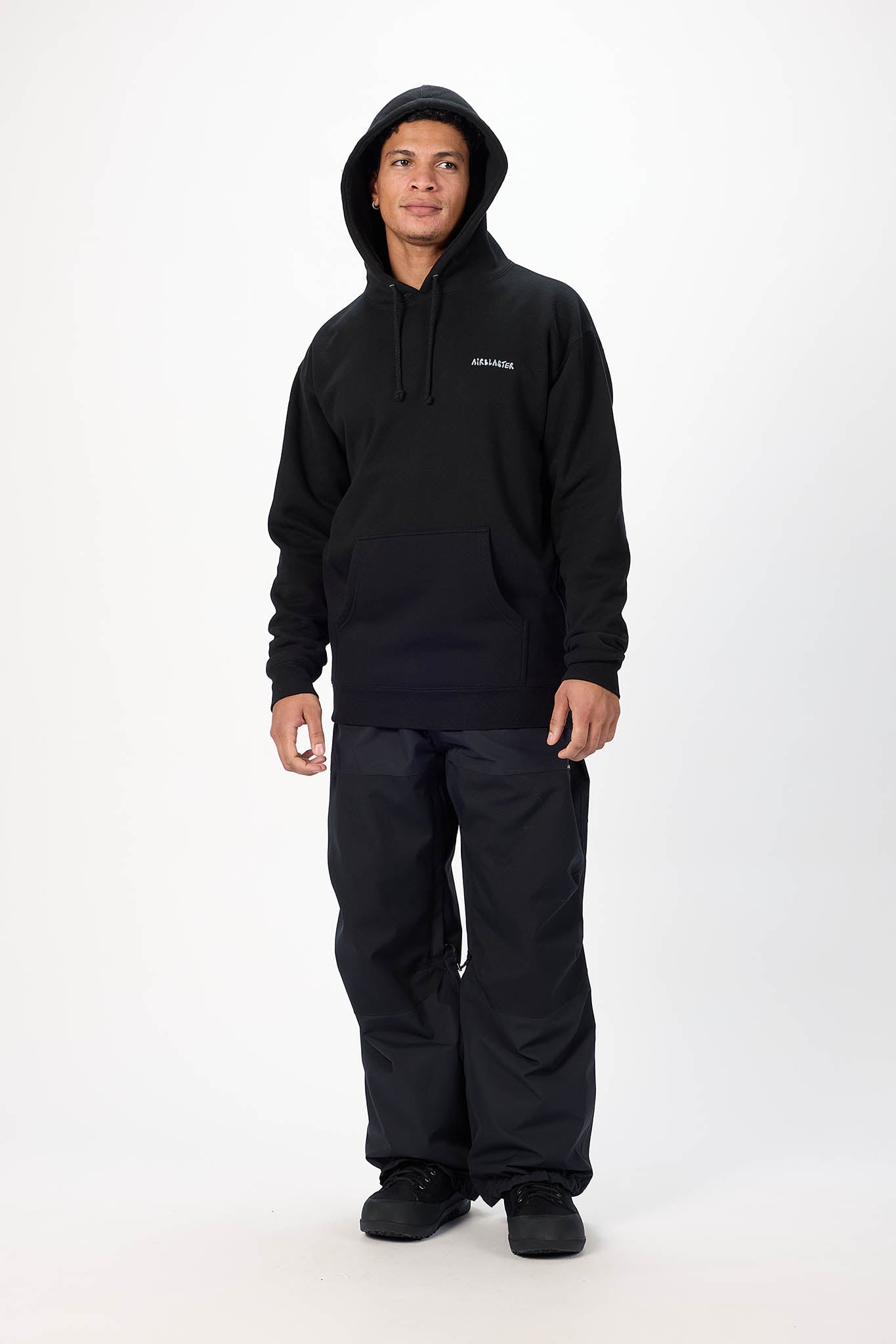 A man wearing a black hooded sweatshirt and black pants, standing against a plain white studio background. A small white logo is visible on the left chest of the hoodie.