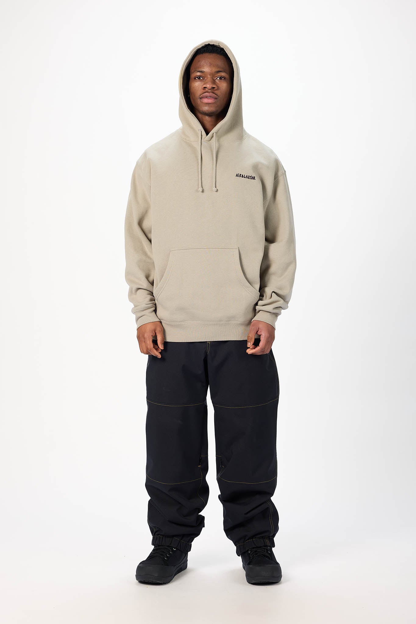 A model wearing a beige pullover hoodie with a small black logo on the left chest, paired with black pants and black sneakers, standing against a plain white studio background.