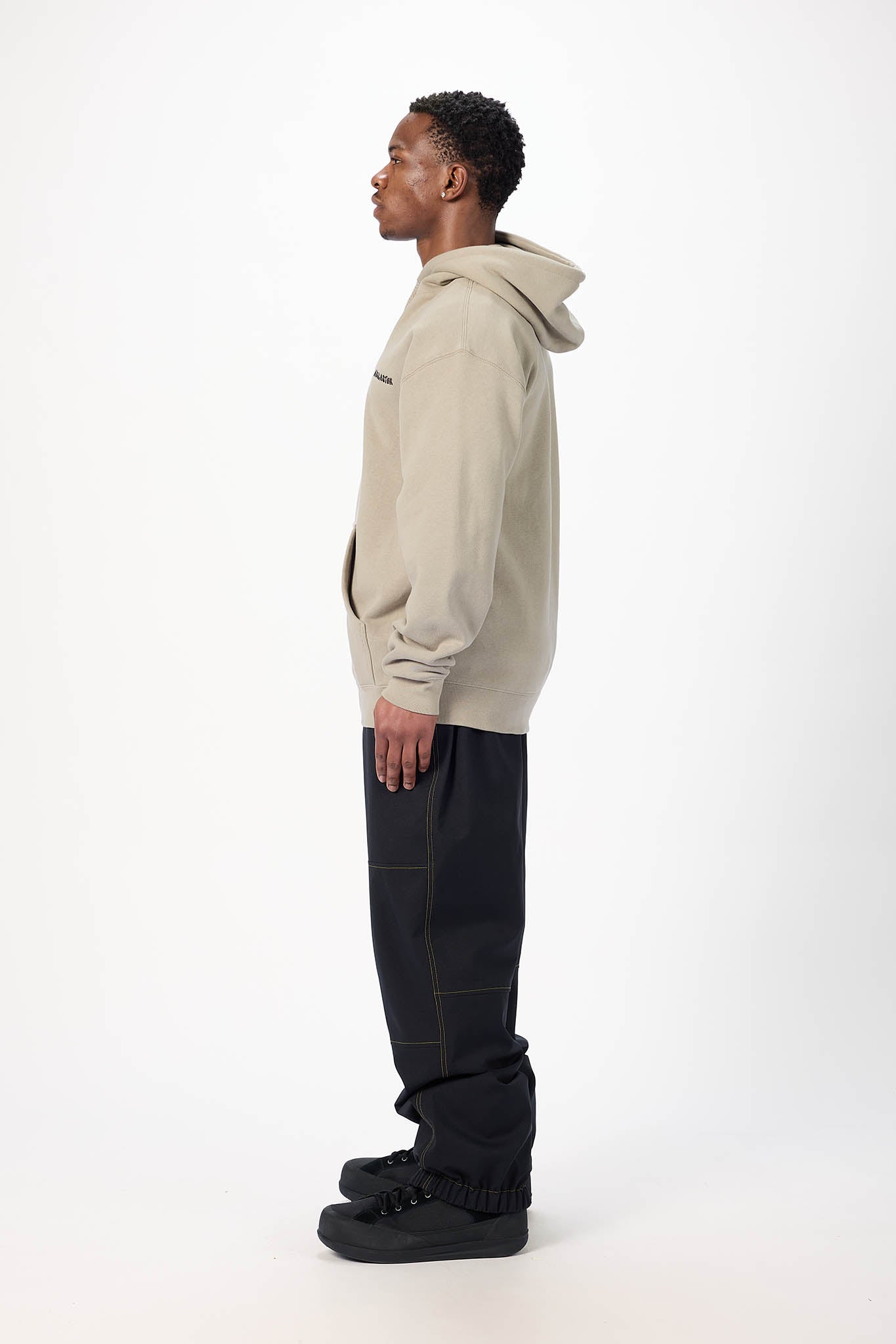 A man in a side profile wearing a light beige oversized hoodie and black pants, standing against a plain white studio background.