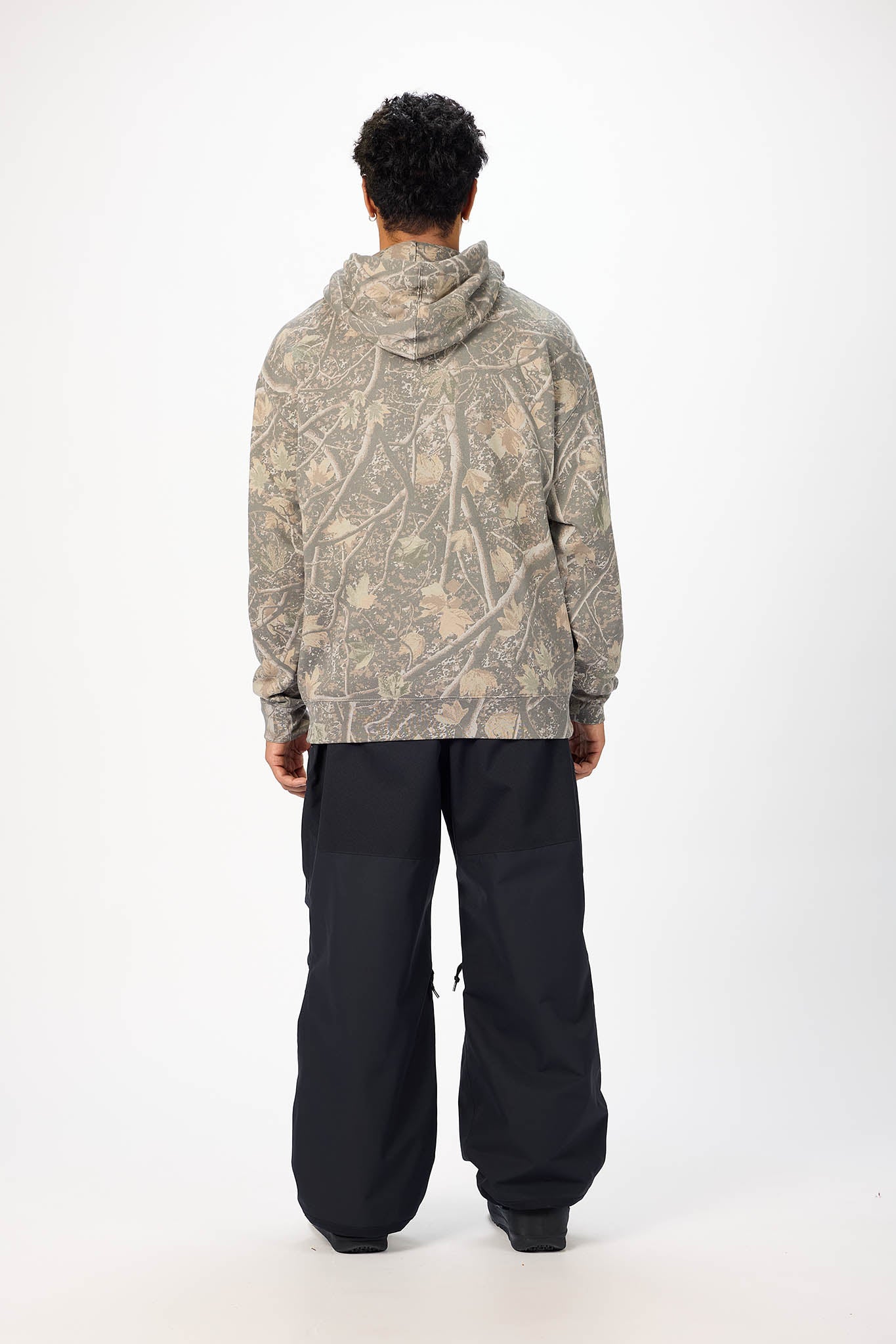 A model viewed from behind wearing a beige and gray camouflage patterned hoodie and black pants, standing against a plain white studio background.