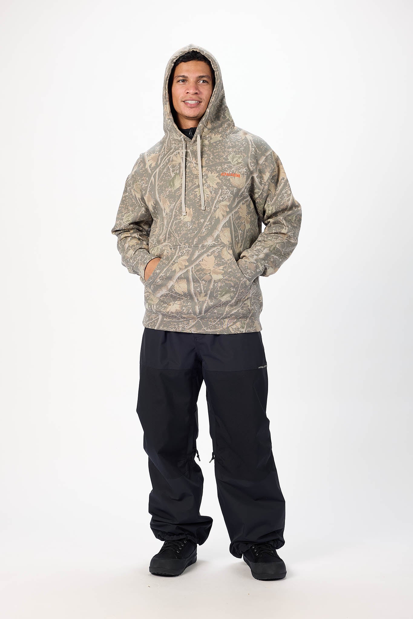 A man wearing a camouflage hoodie with a small orange chest logo and black pants, standing against a plain white studio background.
