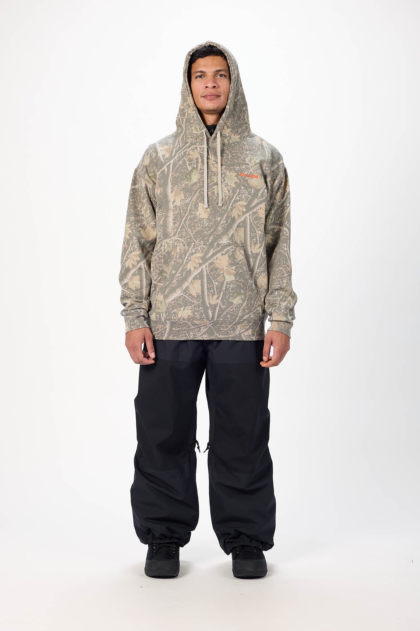 A model stands against a light gray studio background wearing a camouflage-patterned hooded sweatshirt with a small orange chest logo and loose black pants.