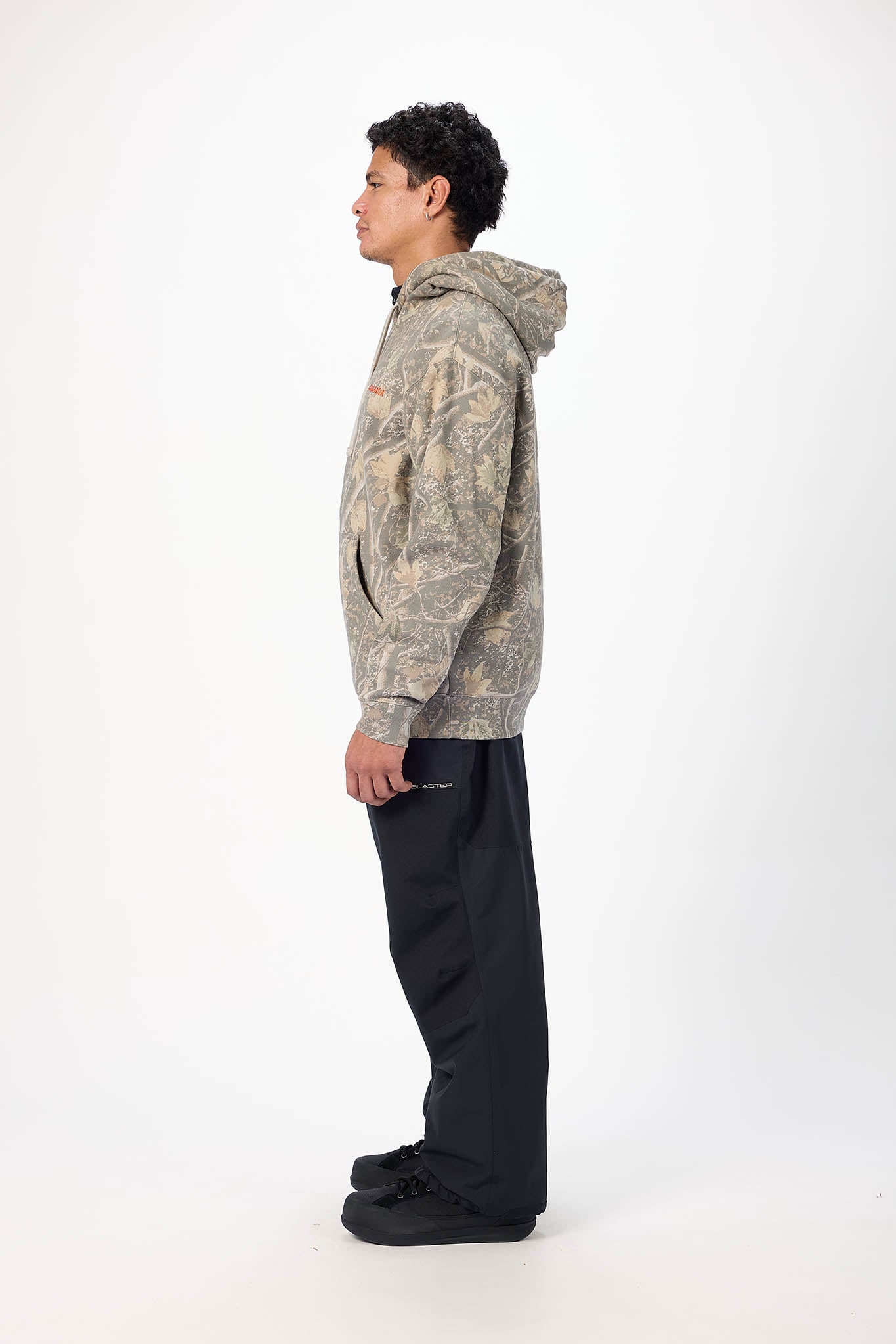 A person standing in profile wearing a camouflage-patterned hoodie and dark pants against a plain light gray studio background.