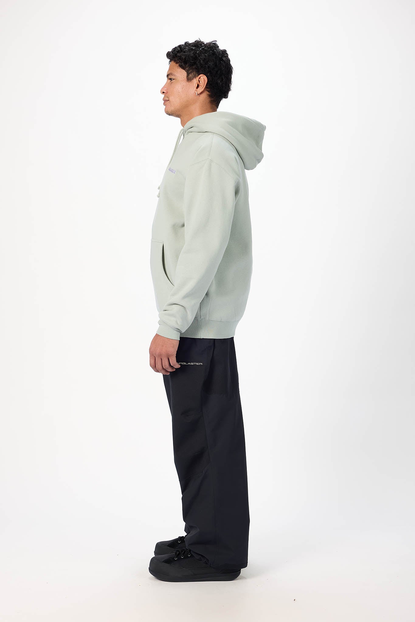 A man in profile wearing a light sage green pullover hoodie and dark charcoal pants, standing against a clean white studio background.