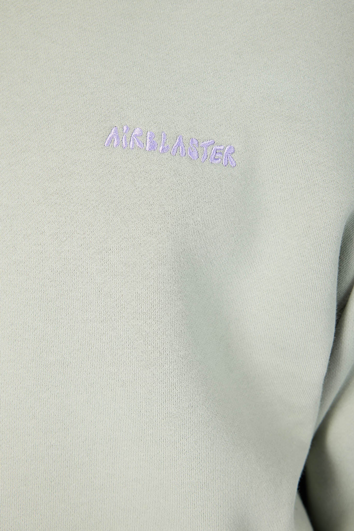 Close-up of a light sage green or gray-green fabric with a small embroidered purple 'AIRBLASTER' logo on the chest.