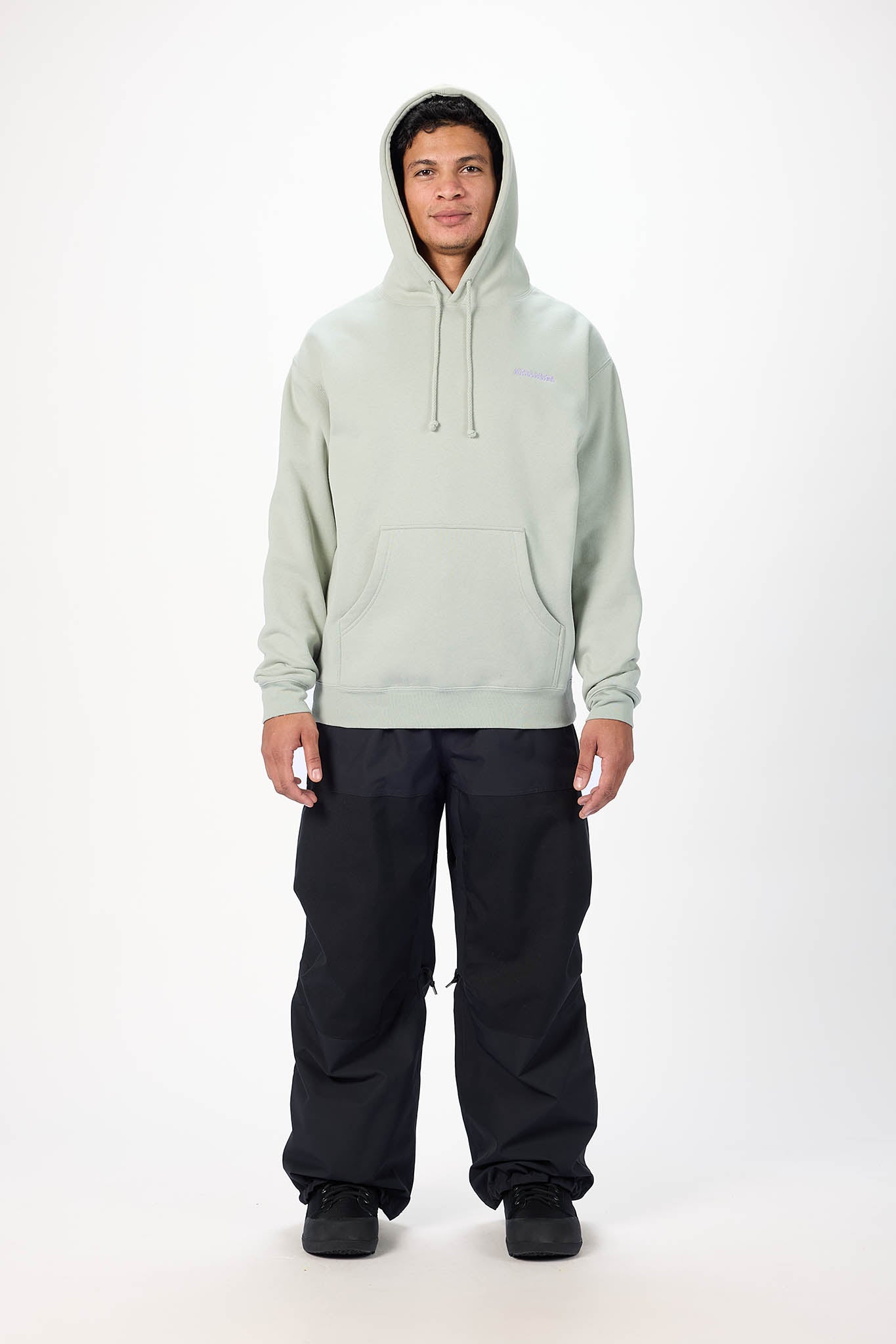 A man wearing a light sage green pullover hoodie with a kangaroo pocket and black pants, standing against a plain white studio background.