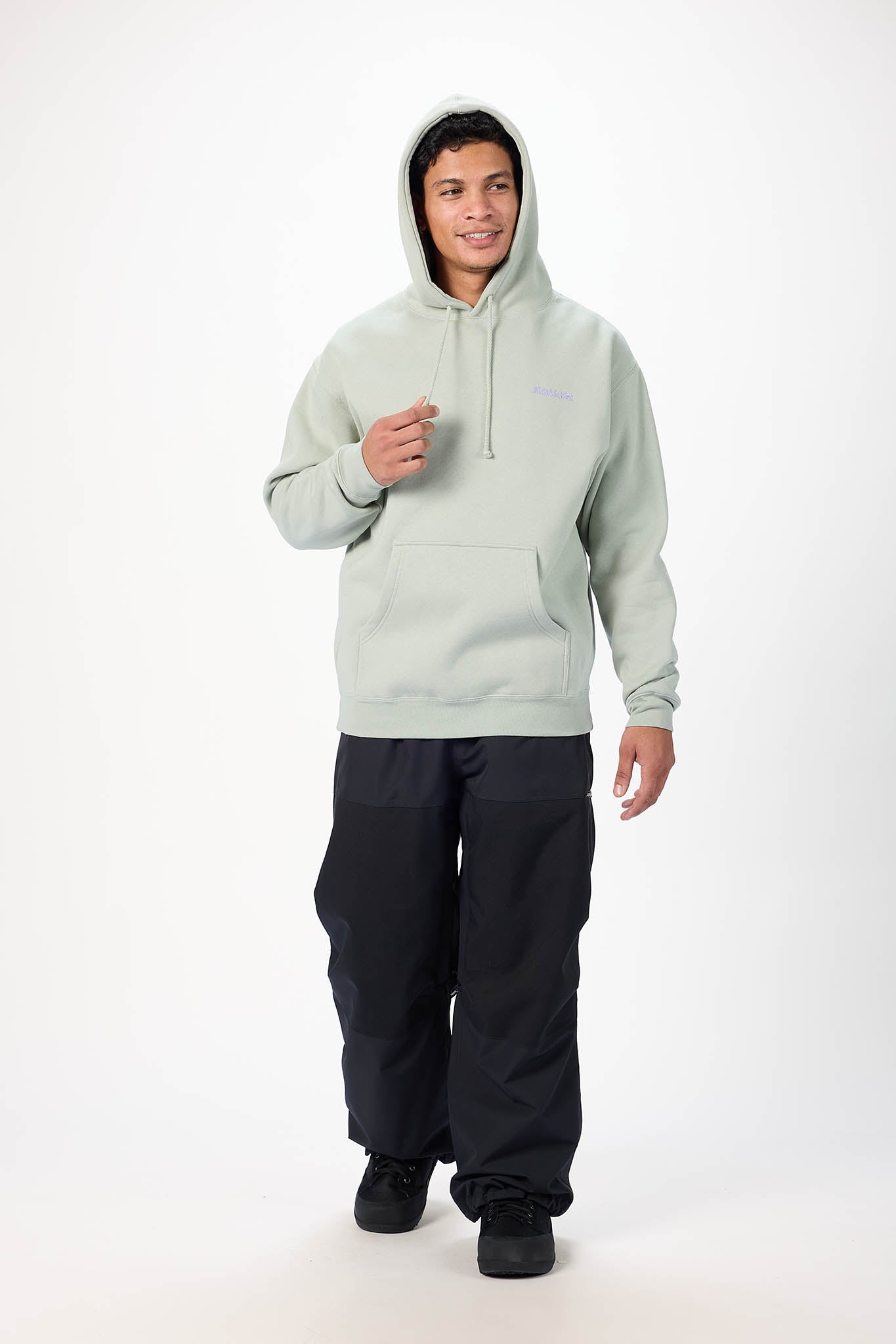 A man wearing a light sage green pullover hoodie with a kangaroo pocket and black pants, standing against a plain white studio background.