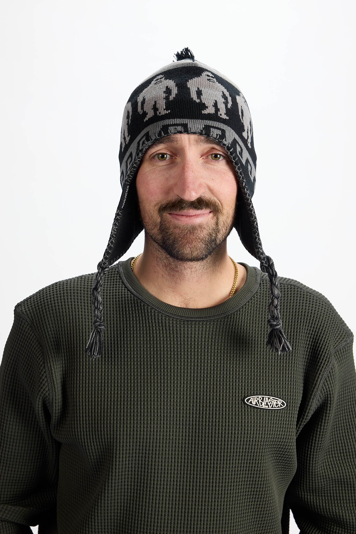 A man wearing a dark green waffle-knit crewneck sweatshirt and a black knit beanie with gray gorilla silhouettes and ear flaps, photographed against a plain white background.