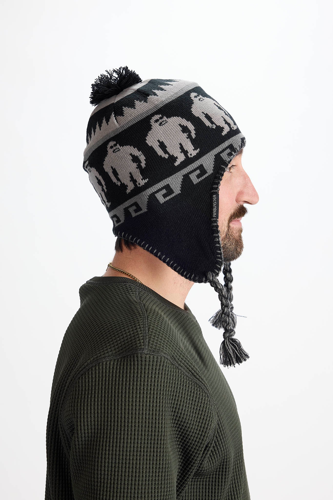 A man shown in profile wearing a black knit winter hat with ear flaps, a black-and-gray patterned band, a black pom-pom, and braided tassels. The background is a clean white studio backdrop.