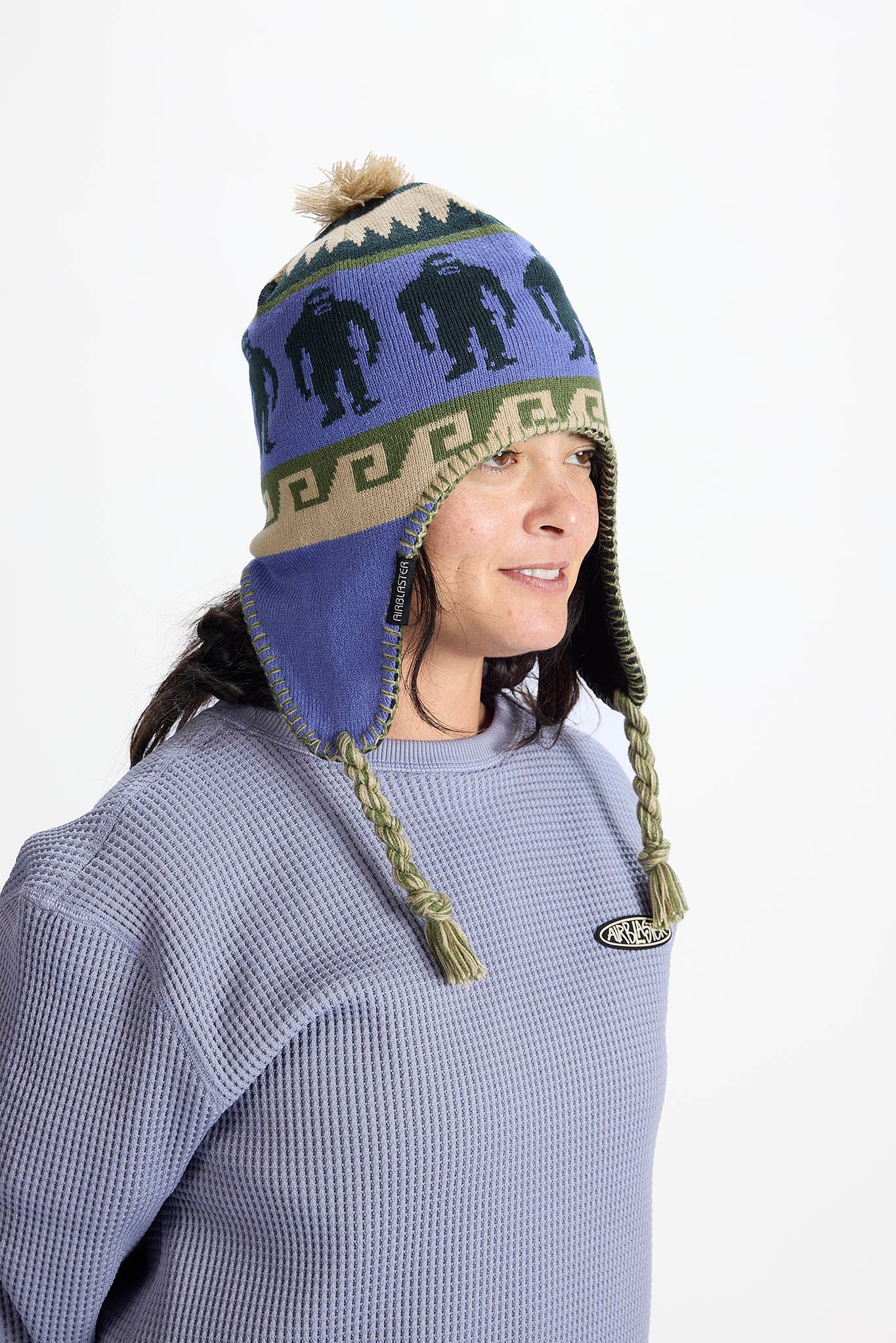 A woman wearing a blue knit winter hat with ear flaps, braided tassels, a tan band, and dark blue gorilla graphics, paired with a matching blue waffle-knit sweater against a plain white background.