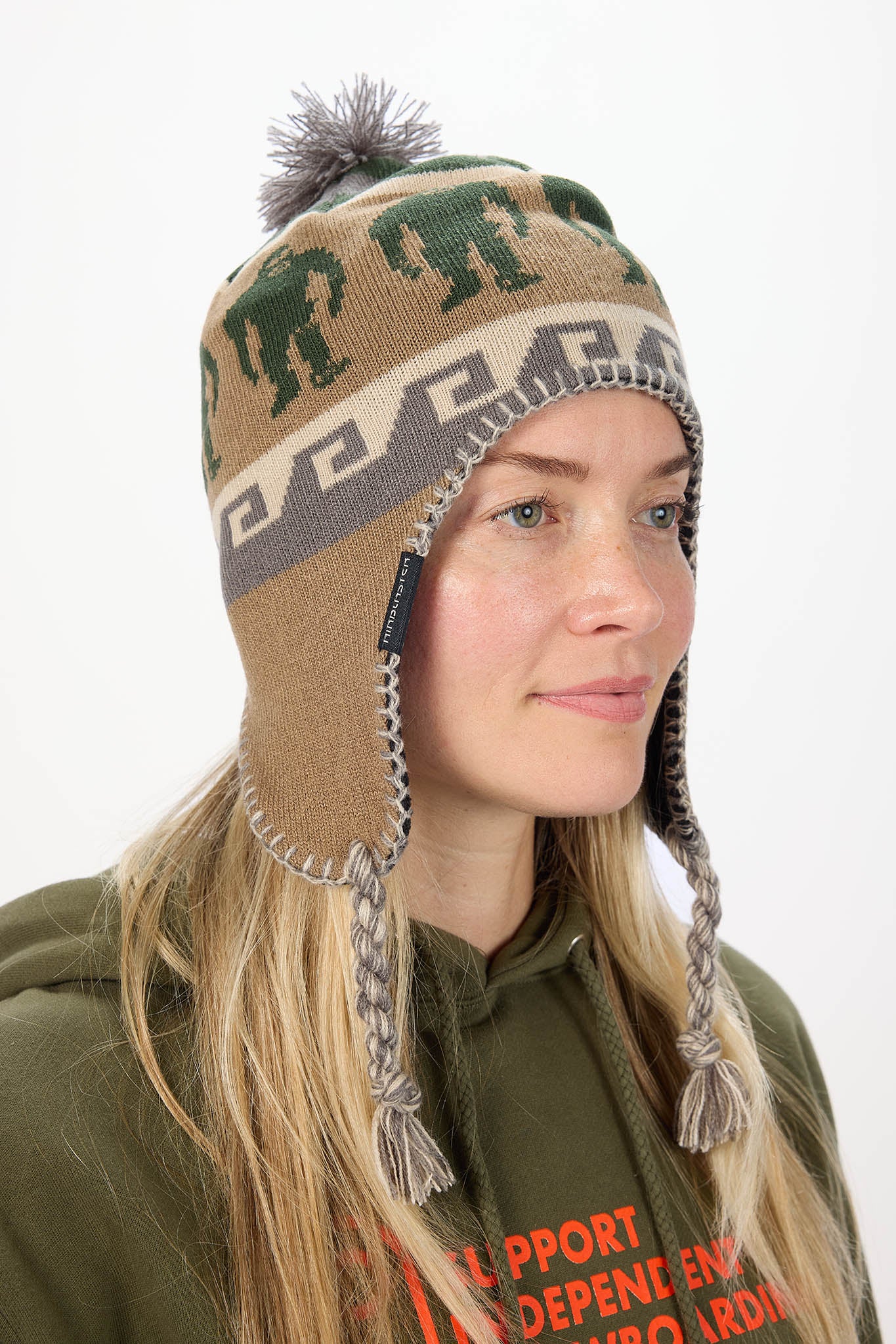 A woman wearing a tan knit winter hat with green elephant motifs, a gray geometric band, braided ear flaps, and a pom-pom on a plain light background.