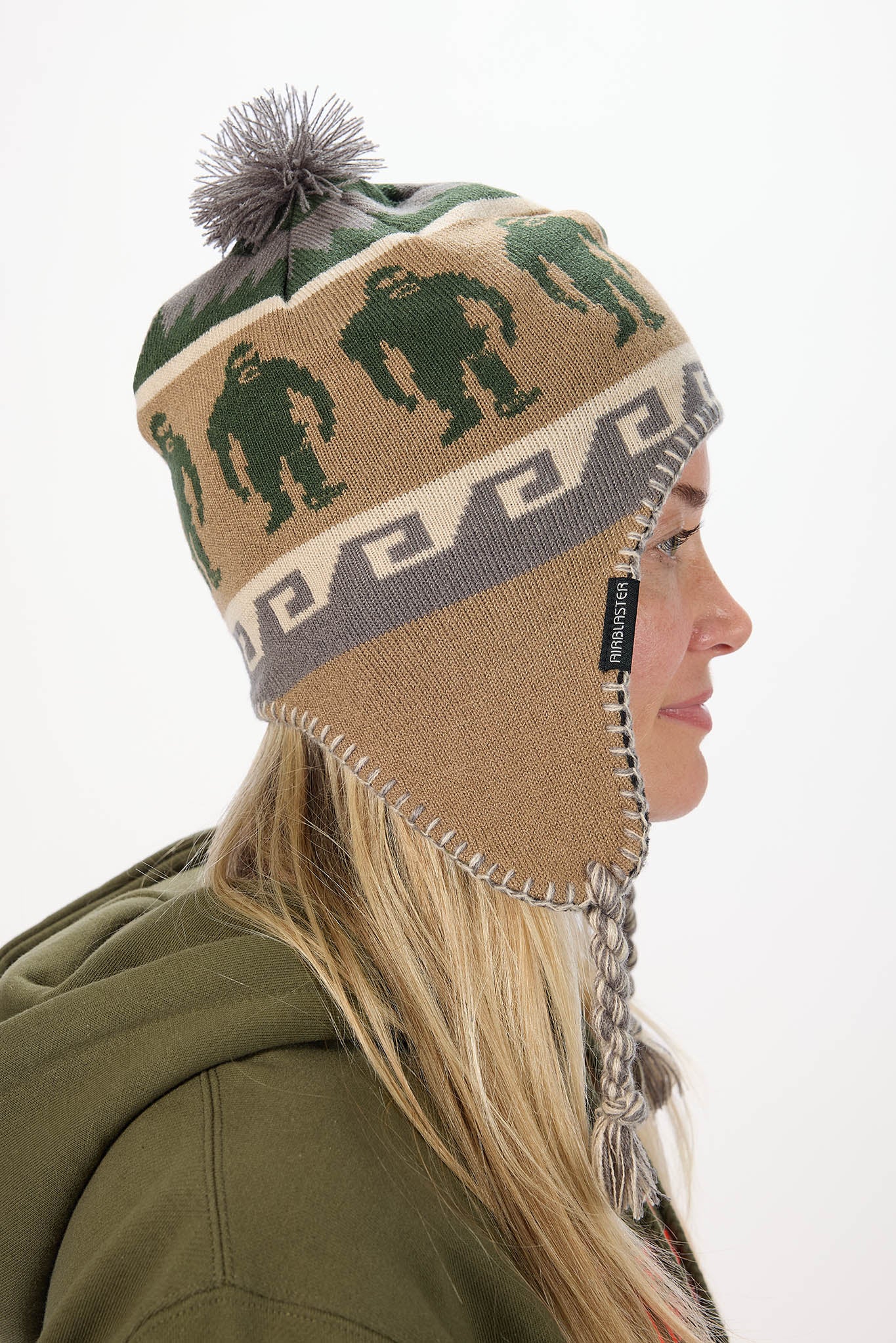 A woman in profile wearing a knit winter hat with a gray pom-pom, green gorilla patterns, and ear flaps, against a plain white background.