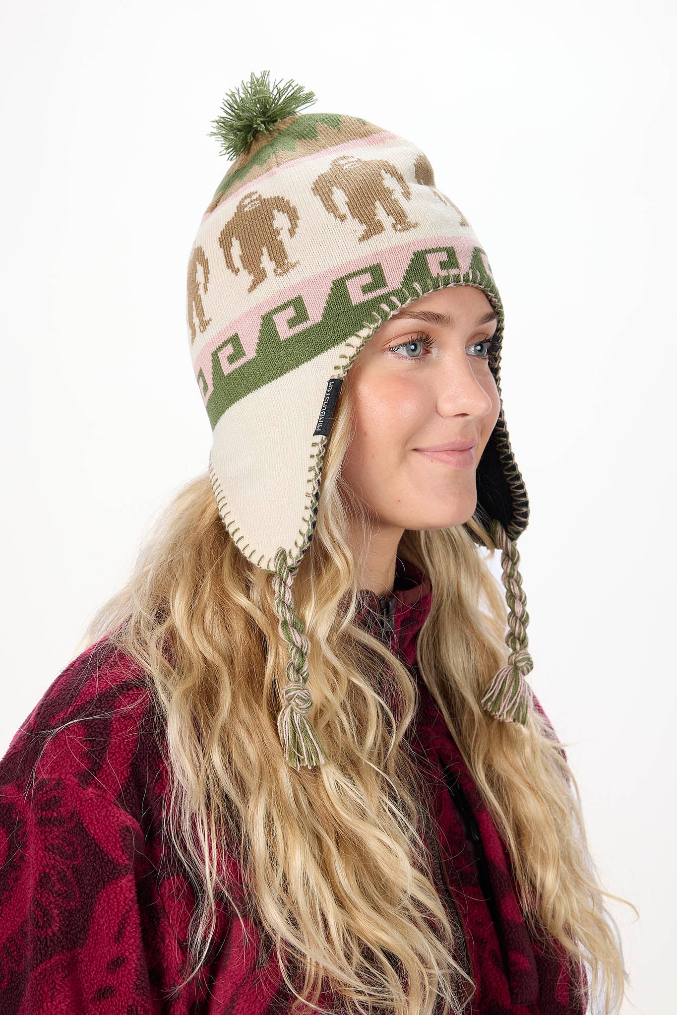 A woman wearing a cream knit winter hat with olive green and burgundy patterns, featuring elephant silhouettes, a pom-pom, and ear flaps with braided ties, set against a plain white background.