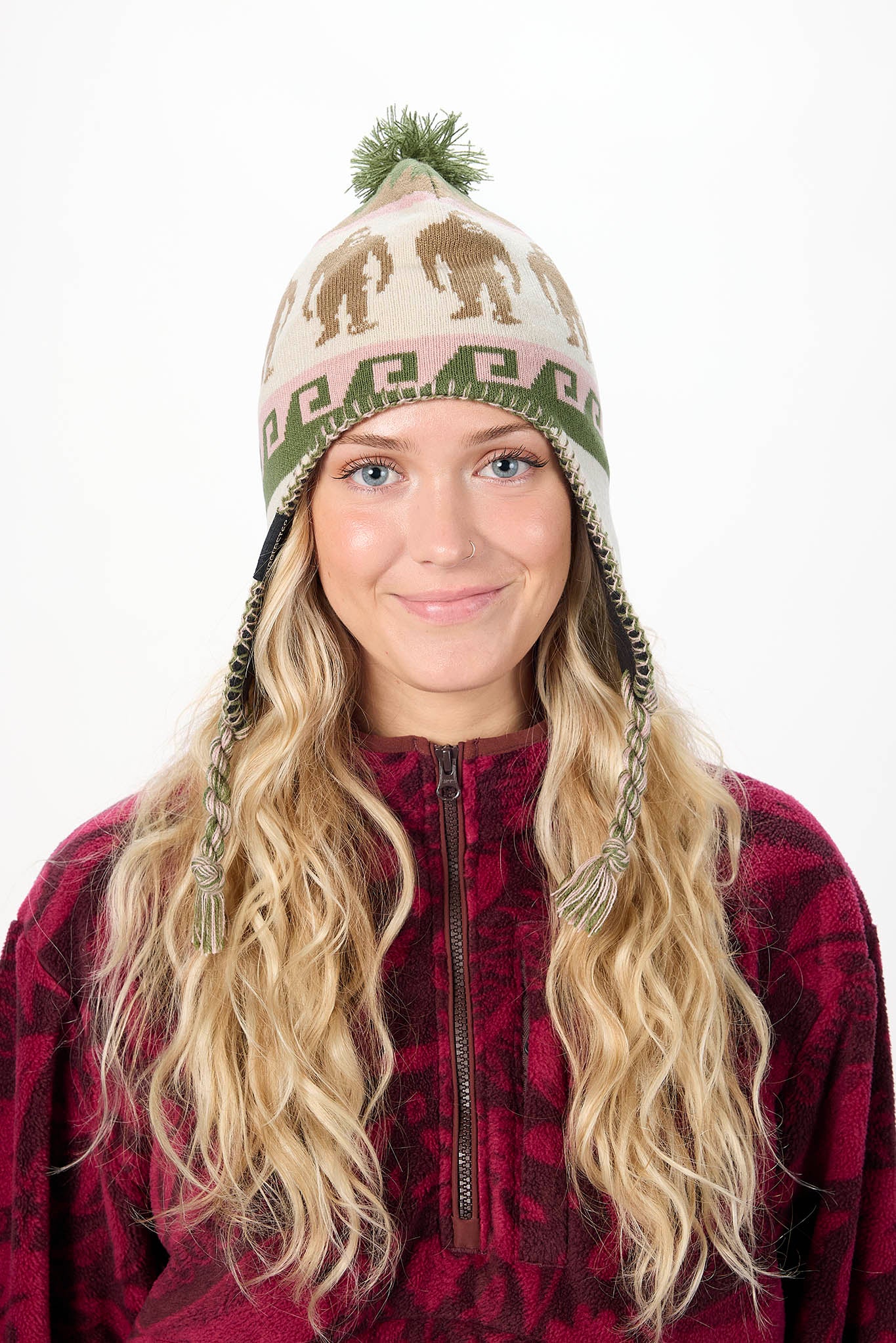 A woman with long blonde hair wearing a knit beanie hat with animal and geometric patterns, paired with a dark red fleece jacket, against a plain white background.