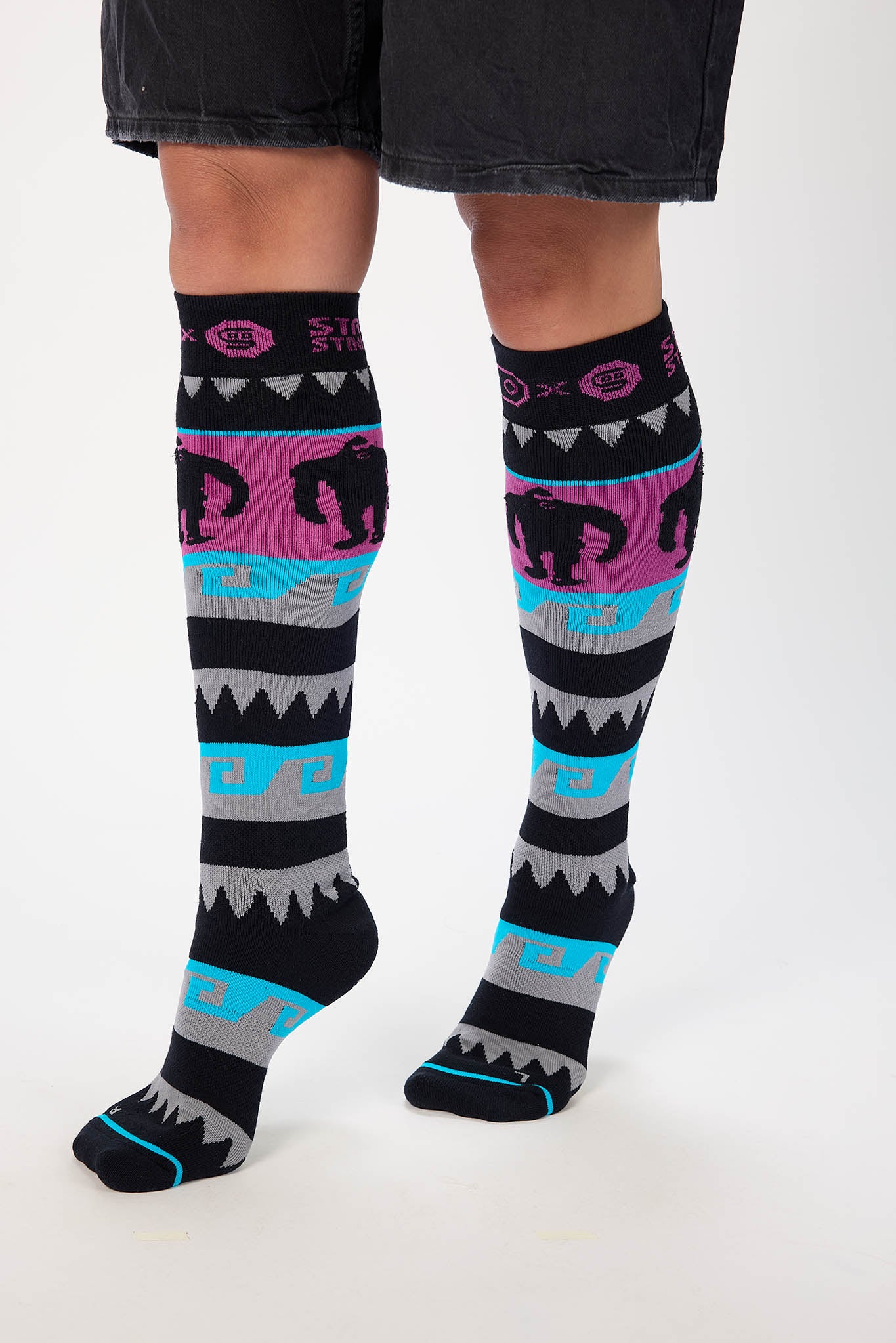 A person wearing patterned knee-high socks with black shorts, shown from the knees down against a plain light background. The socks have a multicolor Southwestern-style design with black, pink, turquoise, gray, and white geometric and animal motifs.