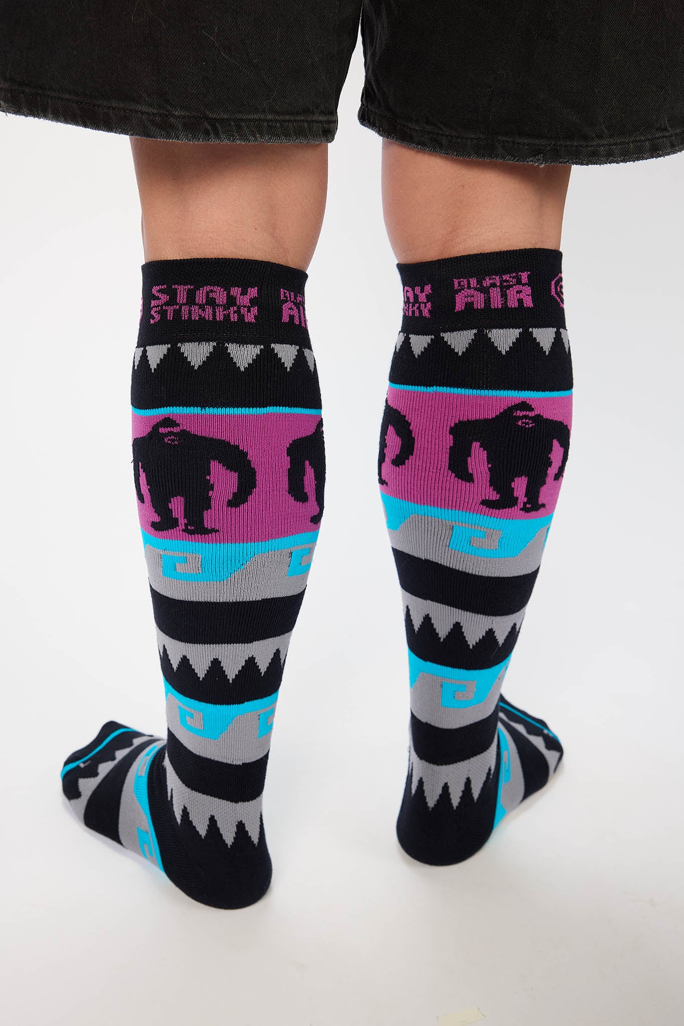 A pair of knee-high socks worn on a person's legs, featuring a black base with a bright pink gorilla pattern, gray and turquoise geometric stripes, and the words 'STAY STINKY' and 'BLAST AIR' on the cuffs.