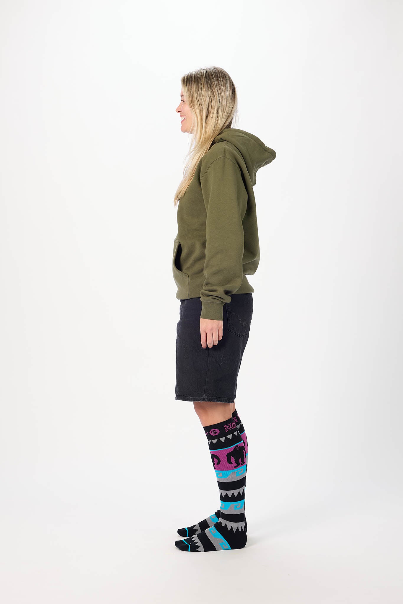 A woman shown in profile wearing an olive green hoodie, a dark navy shorts and black knee-high socks with a colorful geometric and elephant pattern, standing against a plain white studio background.