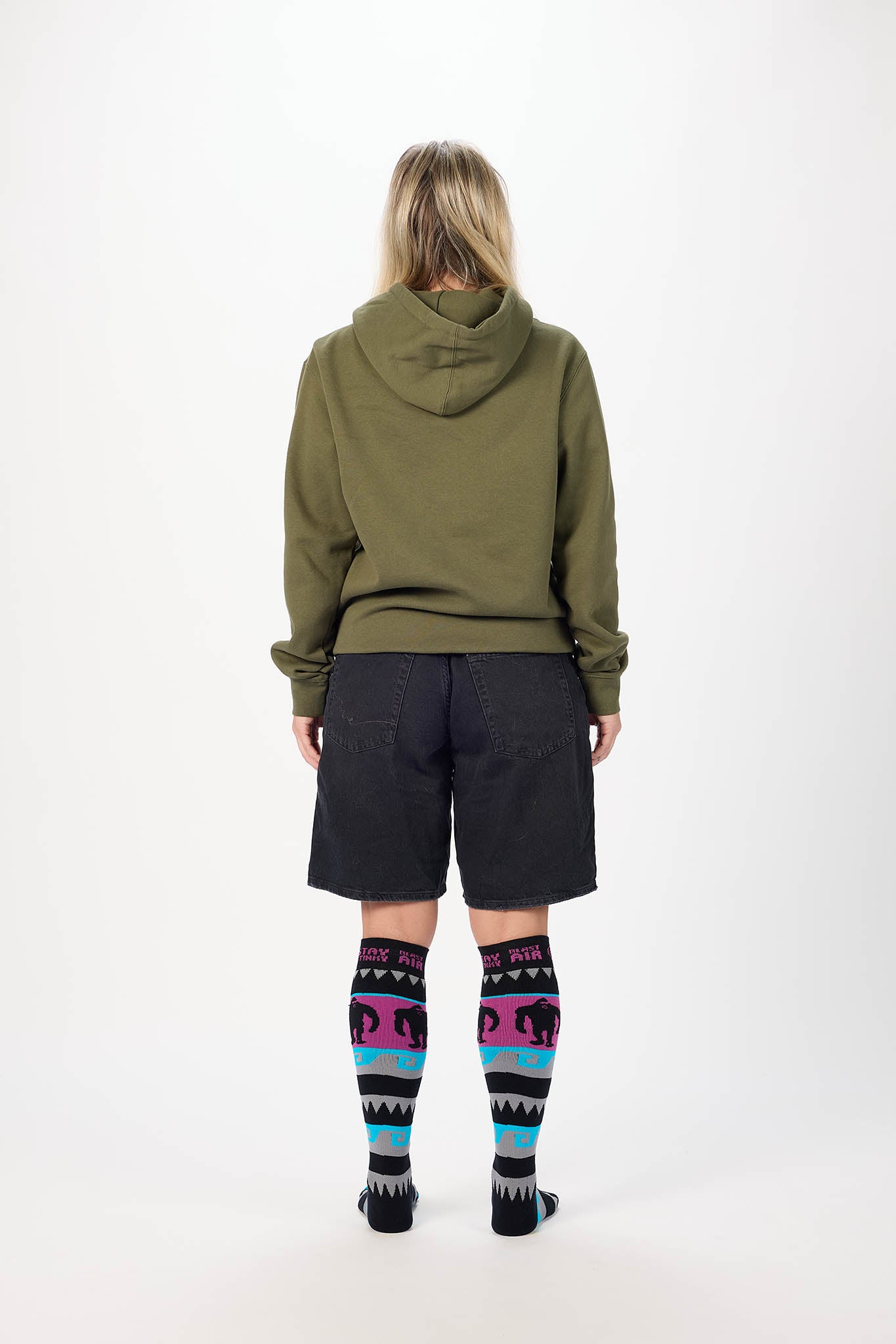A model shown from the back wearing an olive green hoodie, dark charcoal shorts, and black knee-high socks with a colorful geometric pattern in pink, blue, and white.