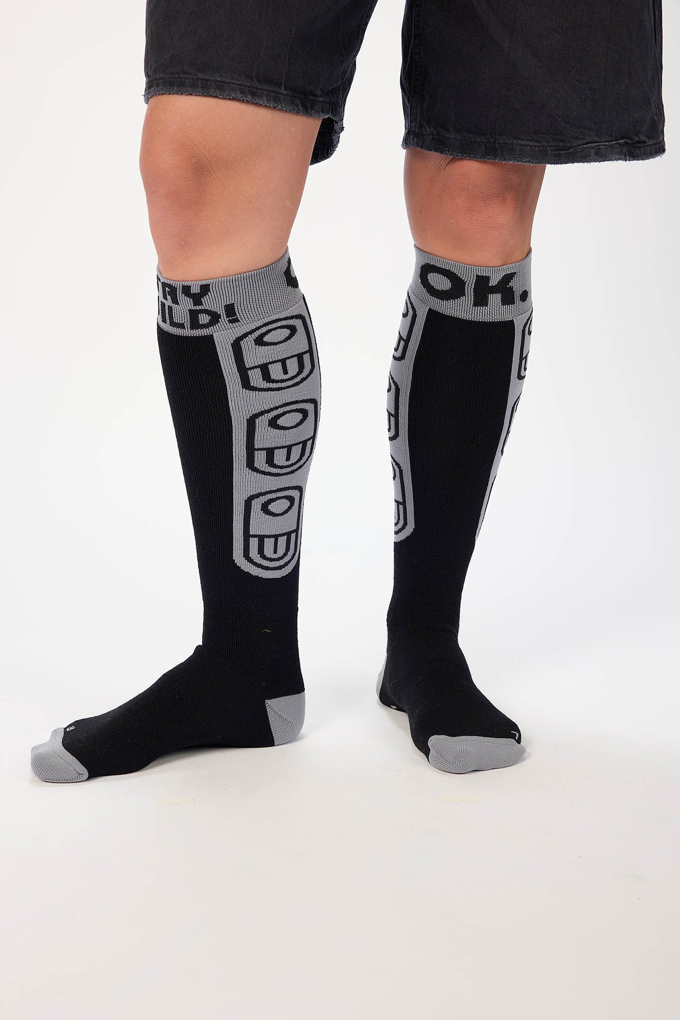 A person wearing black knee-high socks with gray accents and bold graphic text and logo patterns, shown from the knees down against a plain light background.