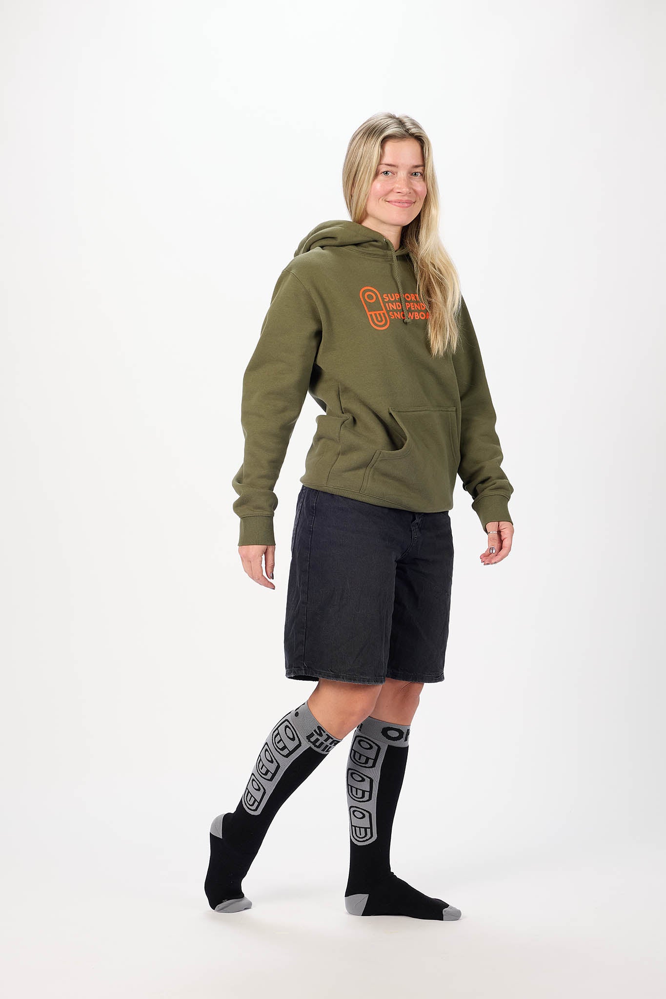 A woman modeling an olive green pullover hoodie with orange embroidered text on the chest, paired with dark shorts and patterned black knee-high socks, against a plain white studio background.