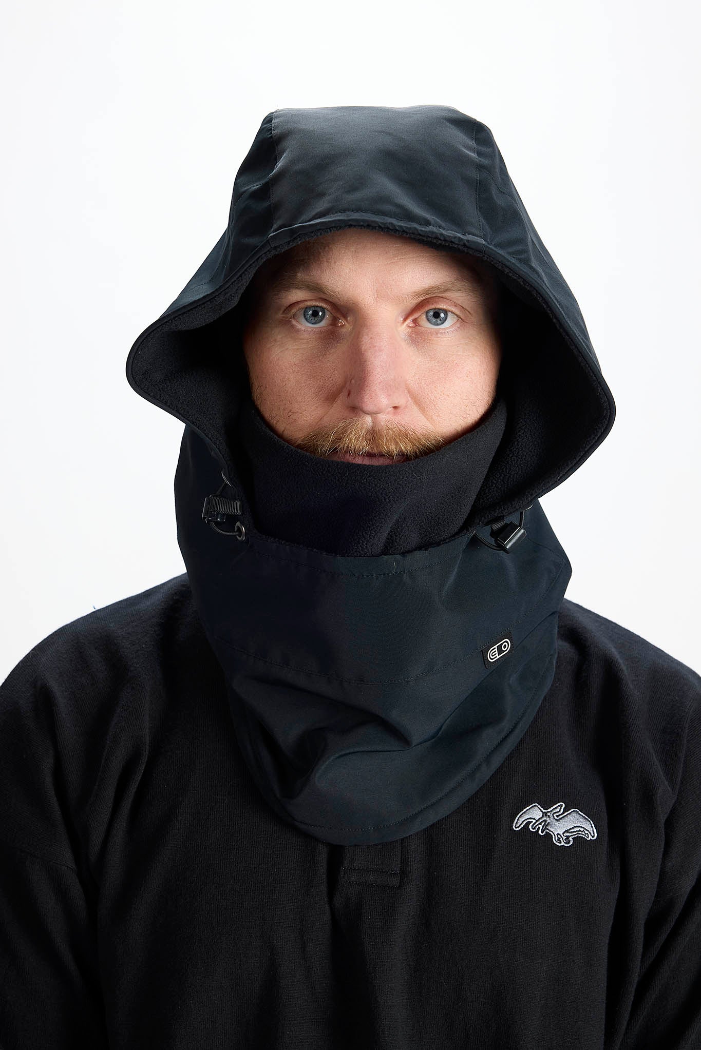 A man wearing a black hooded jacket with an integrated high-neck balaclava-style neck gaiter, shown against a plain white studio background.