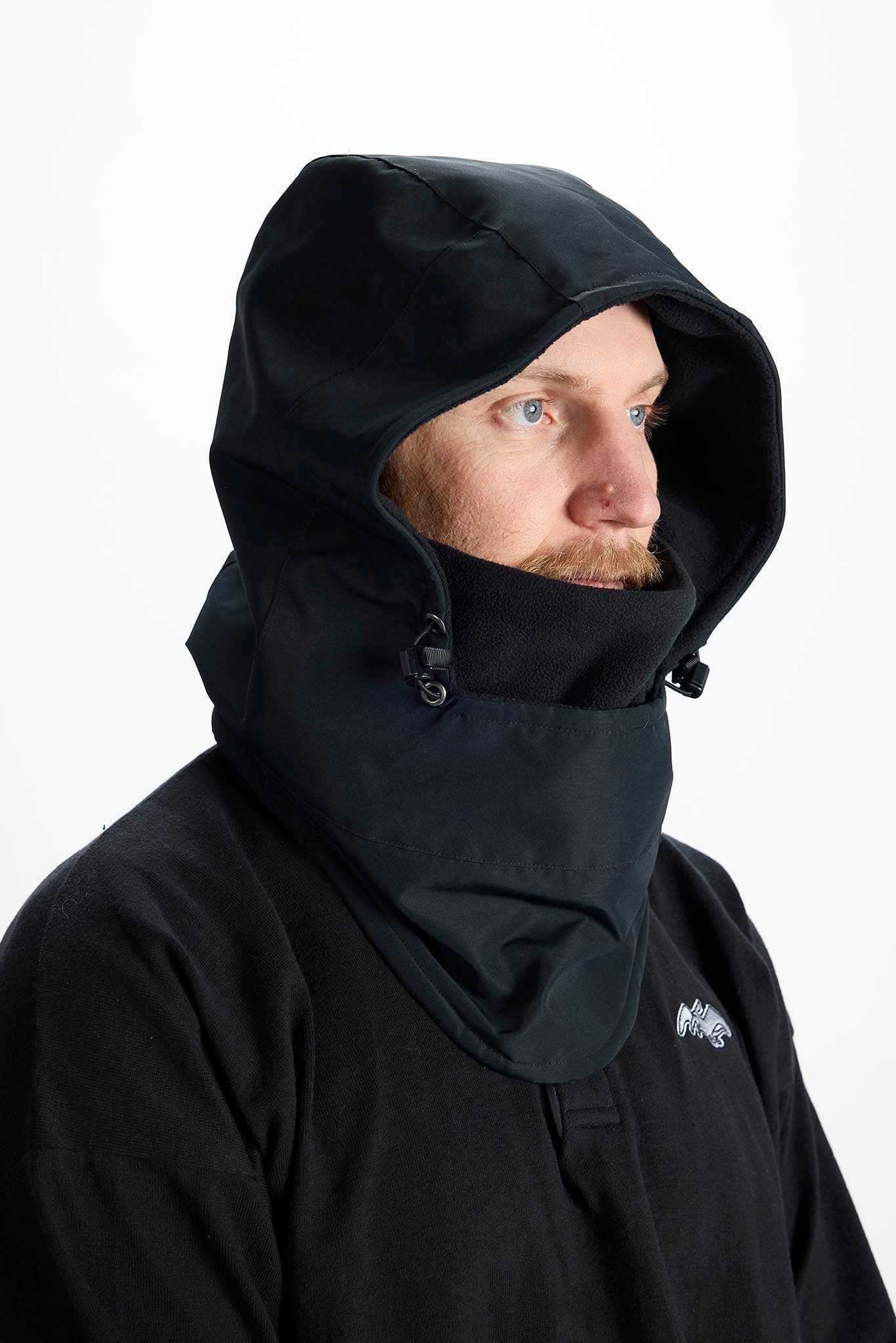 A man wearing a black hooded jacket with an integrated neck gaiter, shown in profile against a plain white studio background.