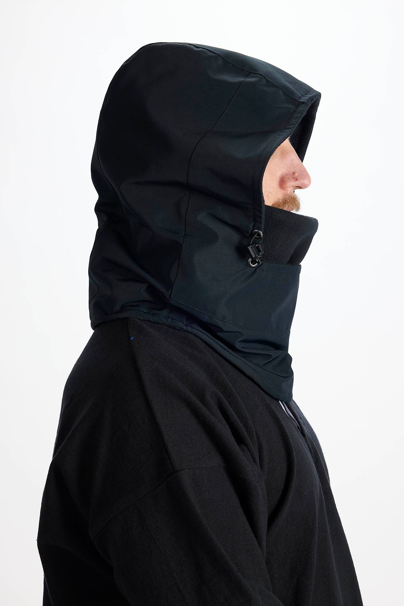 A side profile of a person wearing a black hooded jacket with a high collar and face cover, set against a plain white background.