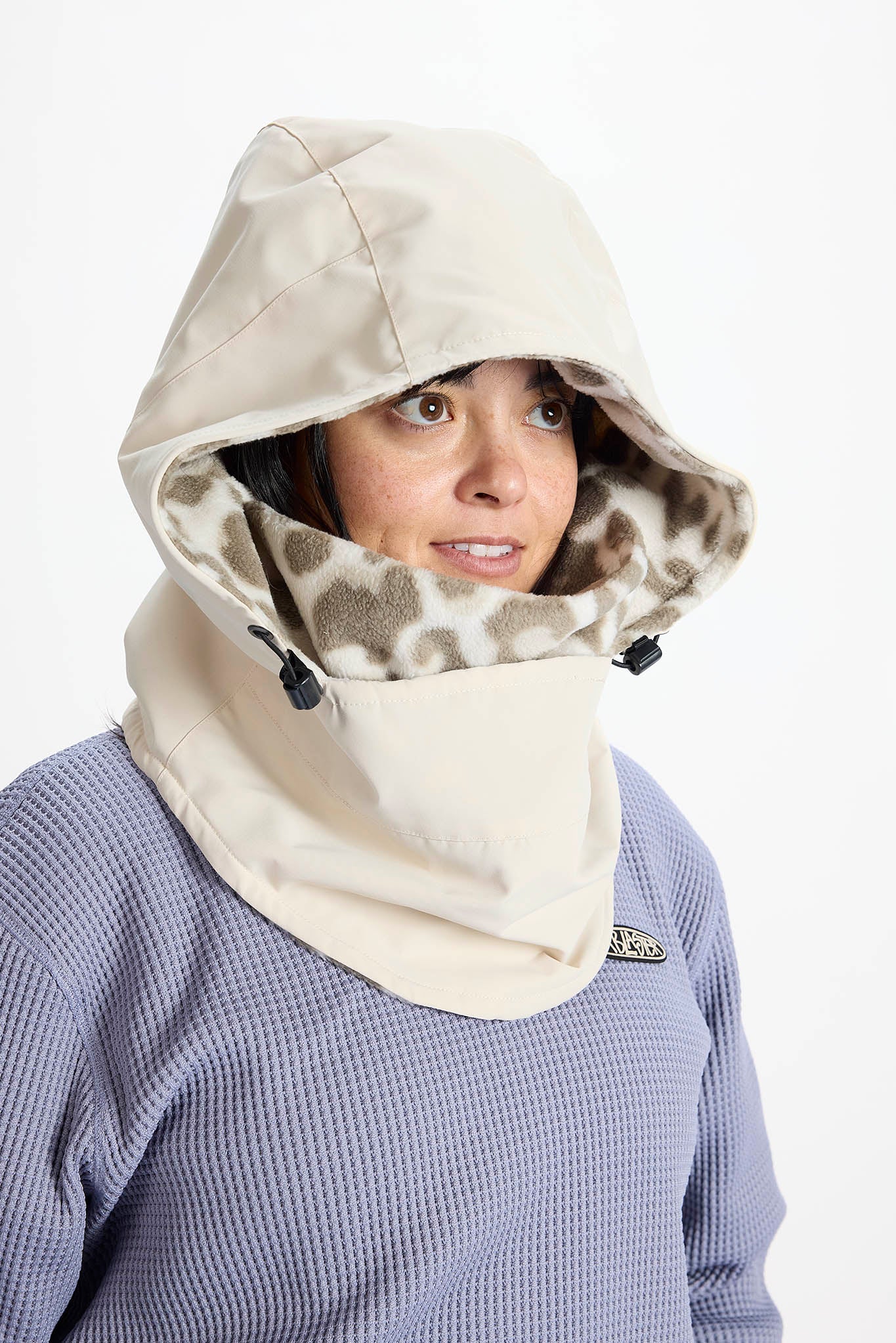 A woman wearing a beige hooded face shield with a leopard-print inner lining, layered over a lavender waffle-knit top, against a plain white background.
