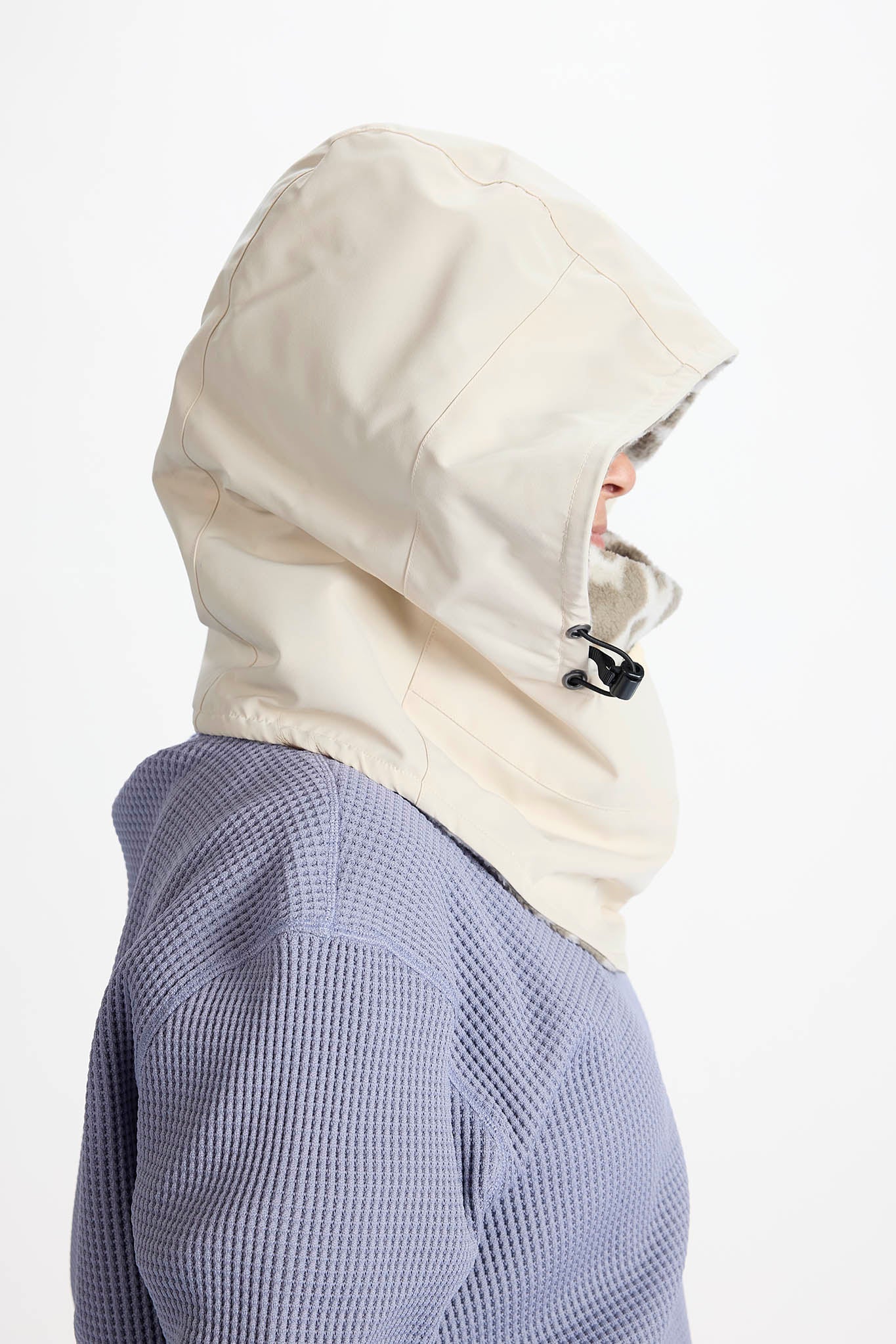 Side profile of a person wearing a light cream hooded face covering or balaclava over a textured periwinkle top, against a plain white background.