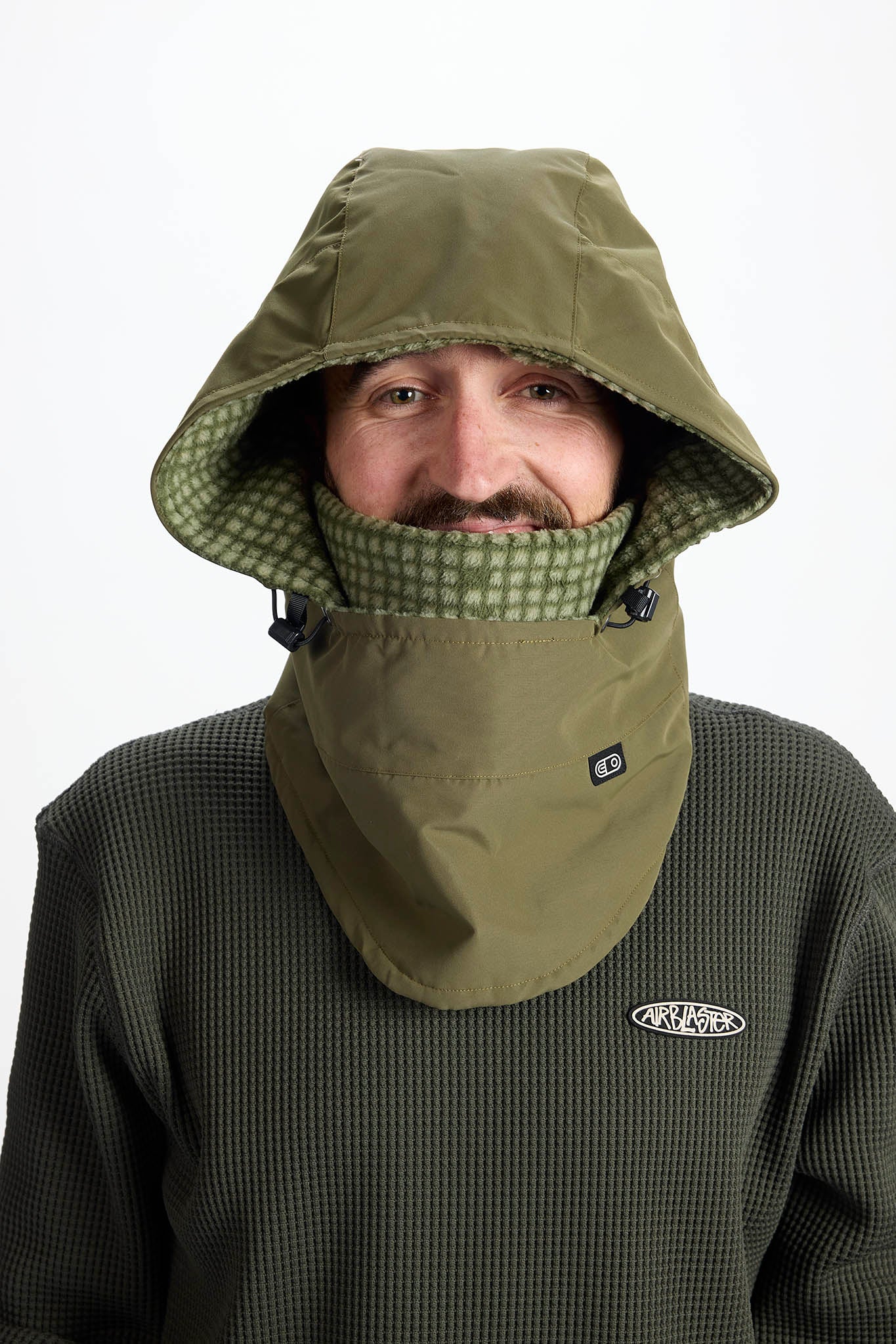 A man wearing an olive green hooded neck warmer and face mask combo over a dark green waffle-knit fleece. The neck warmer features a patterned green and white inner lining and a small white logo on the chest.